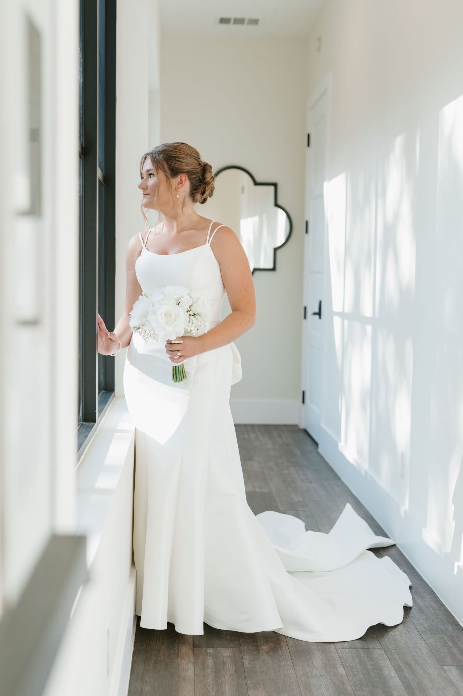 A bride in a flowing white wedding gown stands in an elegant hallway with natural light streaming through windows.
