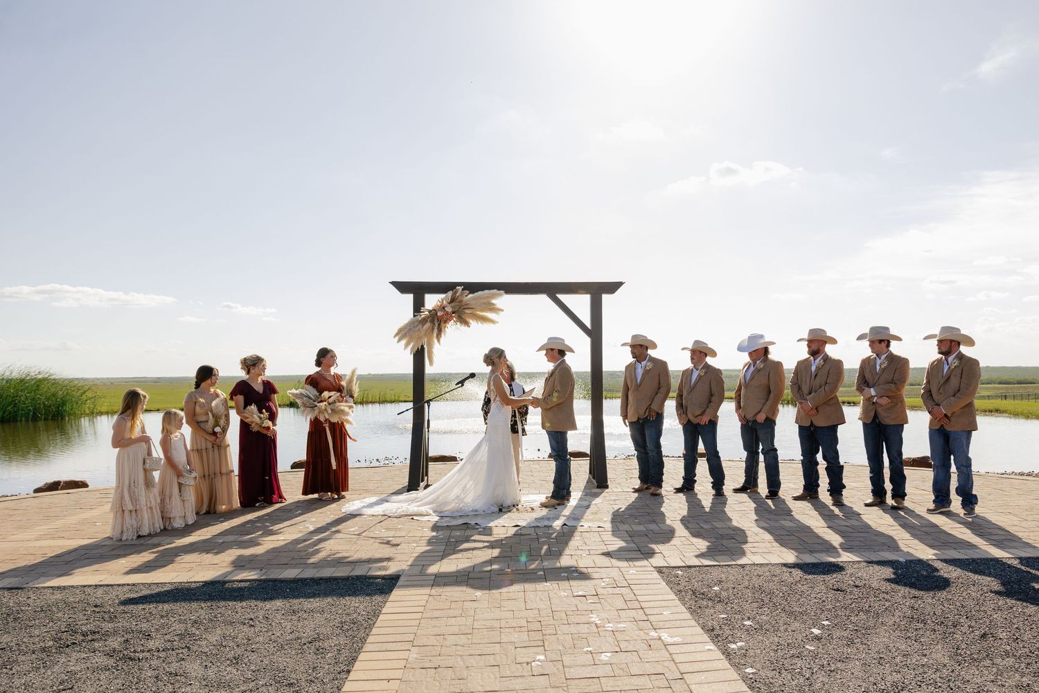 Wedding ceremony group silhouetted against bright sky under wooden arch by waterfront.