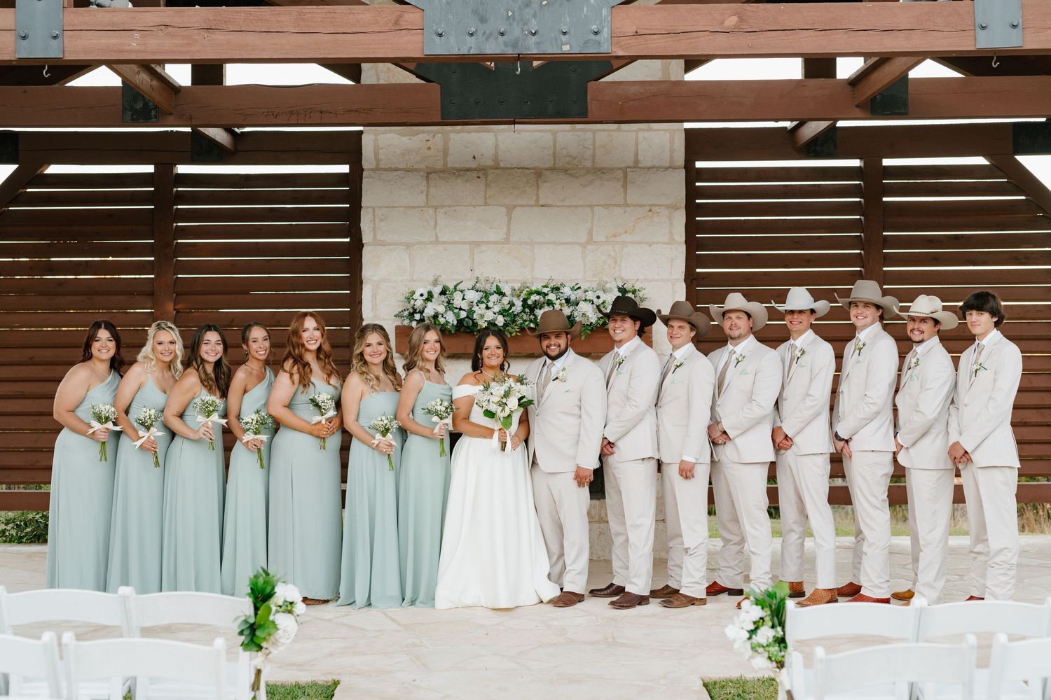 Wedding party in mint green dresses and khaki suits poses together under wooden pergola structure with floral decor.