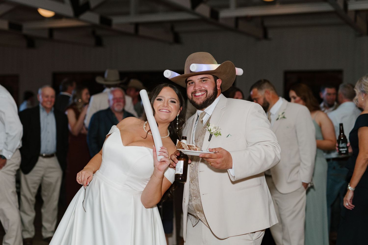 A couple celebrates at a wedding reception indoors, both wearing light colored attire and smiling while holding drinks.