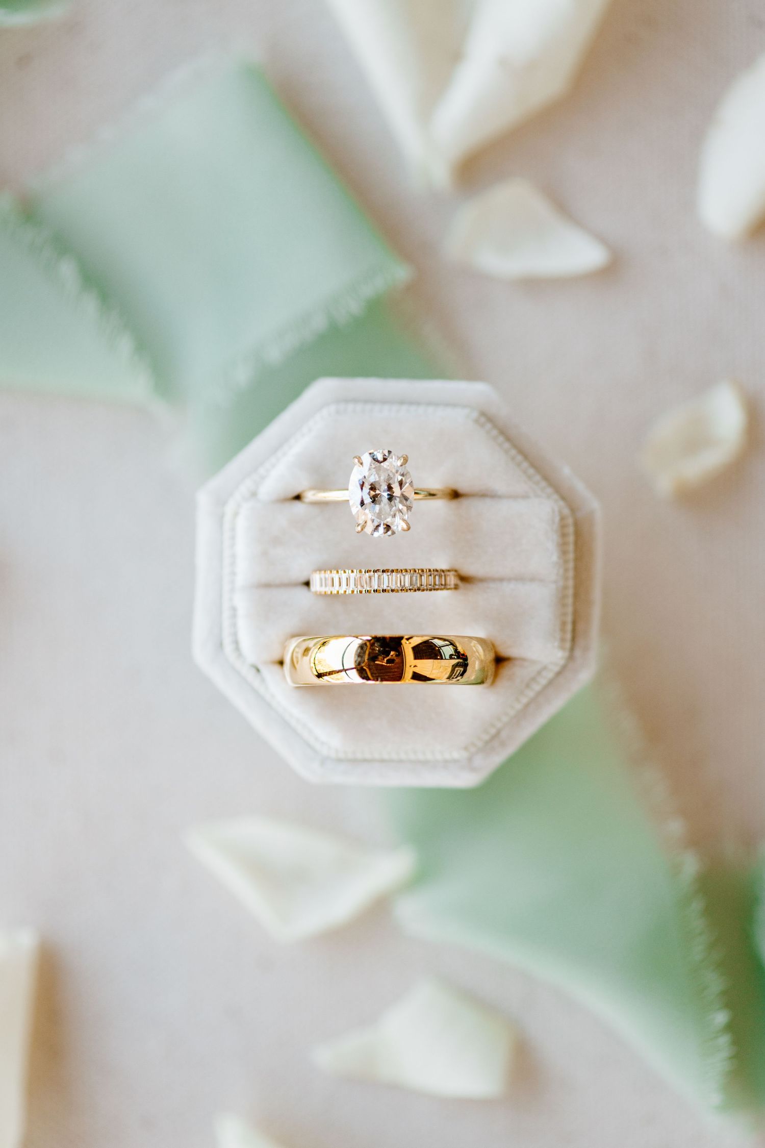 Wedding rings displayed in white octagonal ring box with diamond engagement ring and gold bands against soft pastel background.