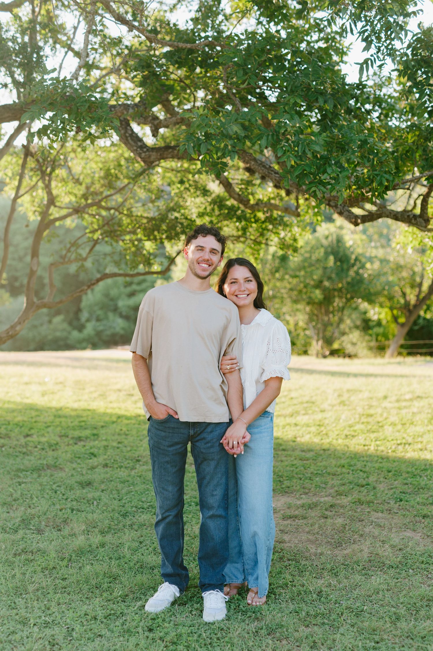 Playful outdoor engagement photos of a couple on grassy hill under tree branches.