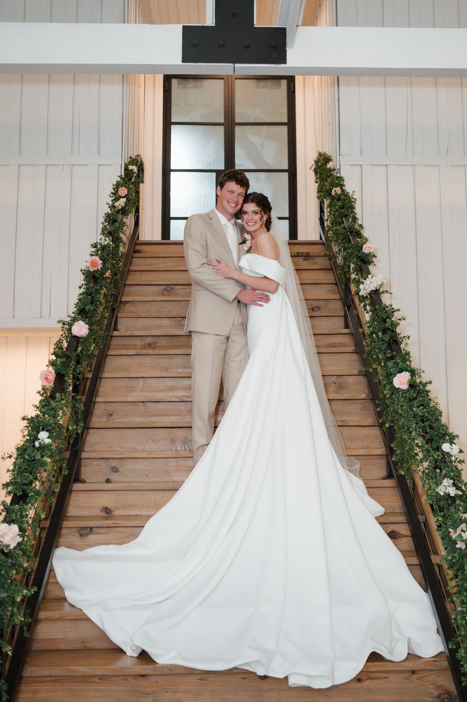 A flowing white wedding dress displayed dramatically on wooden stairs adorned with greenery and floral accents.