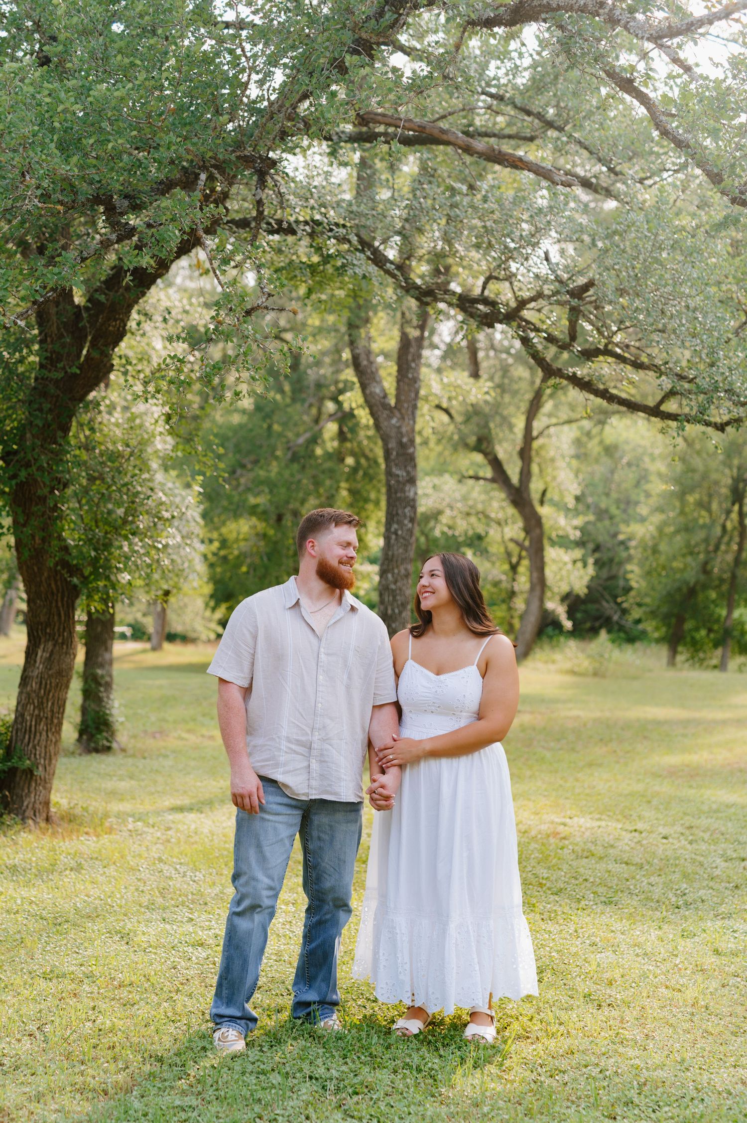 A couple walks hand in hand through a sunlit grove of oak trees in a natural outdoor setting.