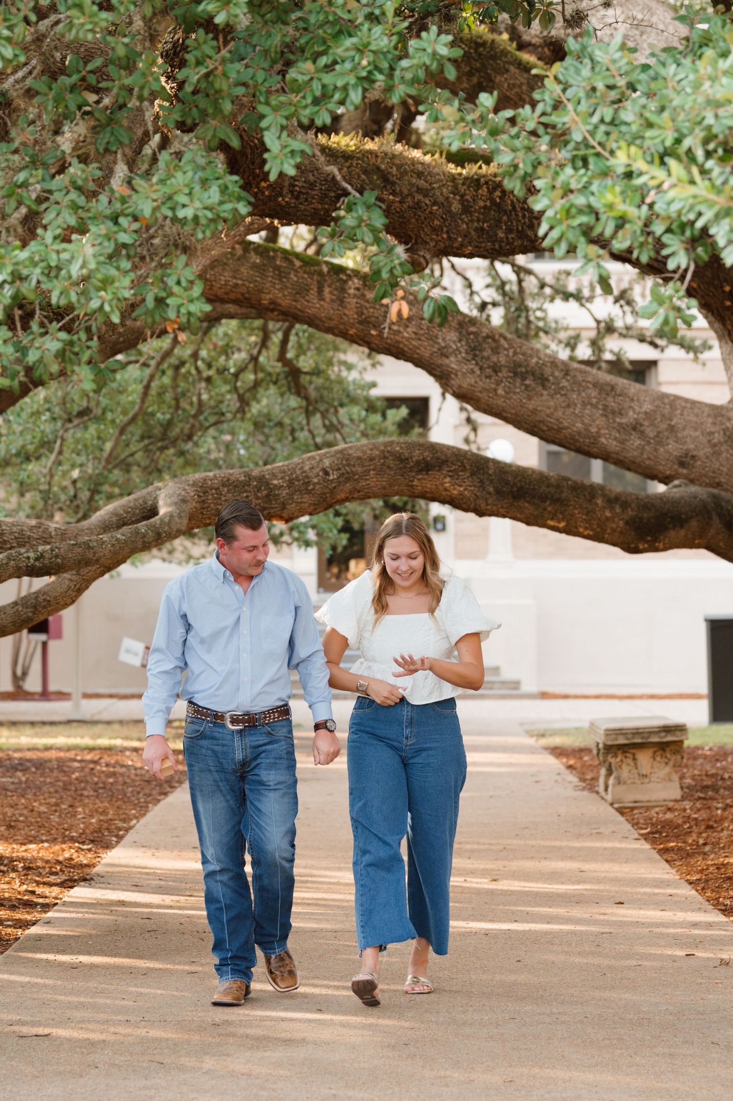 A couple walking beneath a large oak tree branch on a sunny afternoon.
