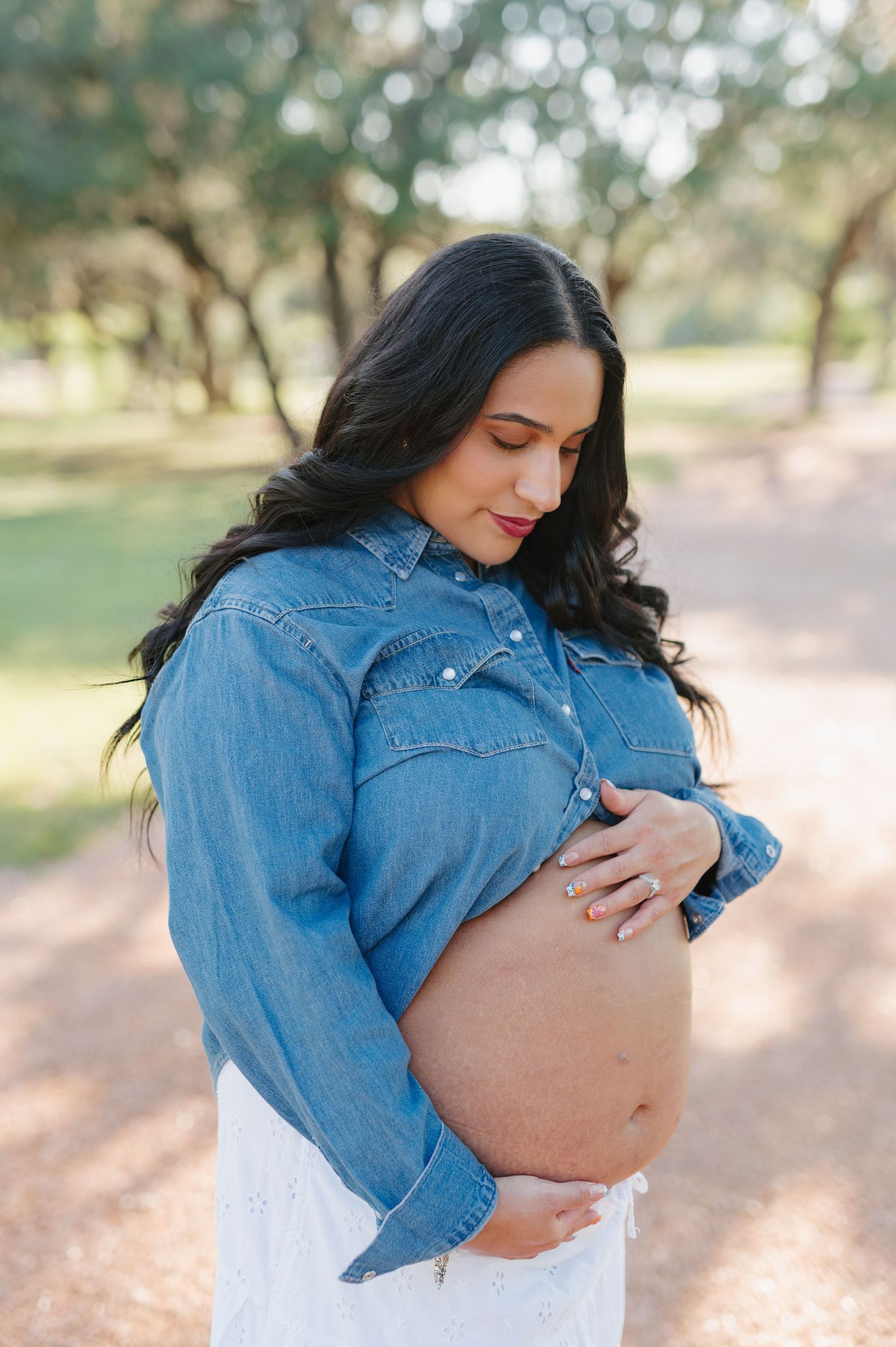 Maternity photos of a woman in denim jacket caressing her baby bump in natural outdoor lighting.