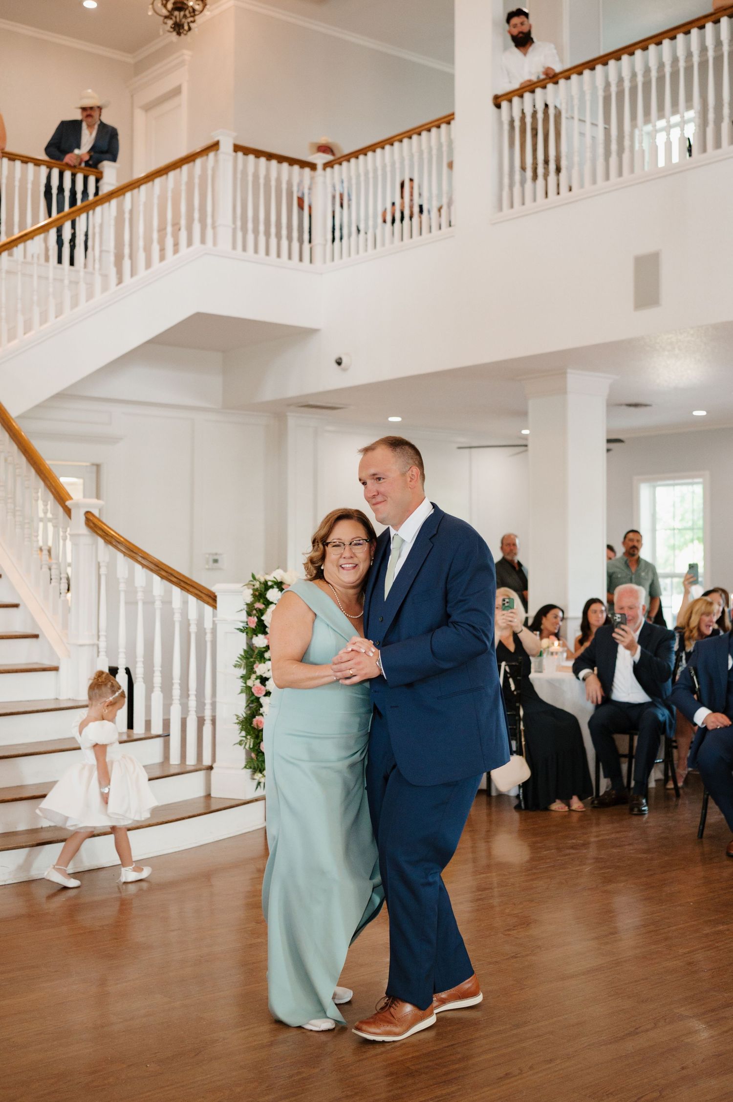 Wedding couple shares a romantic dance together on a wooden floor with white banisters and staircases in elegant venue.