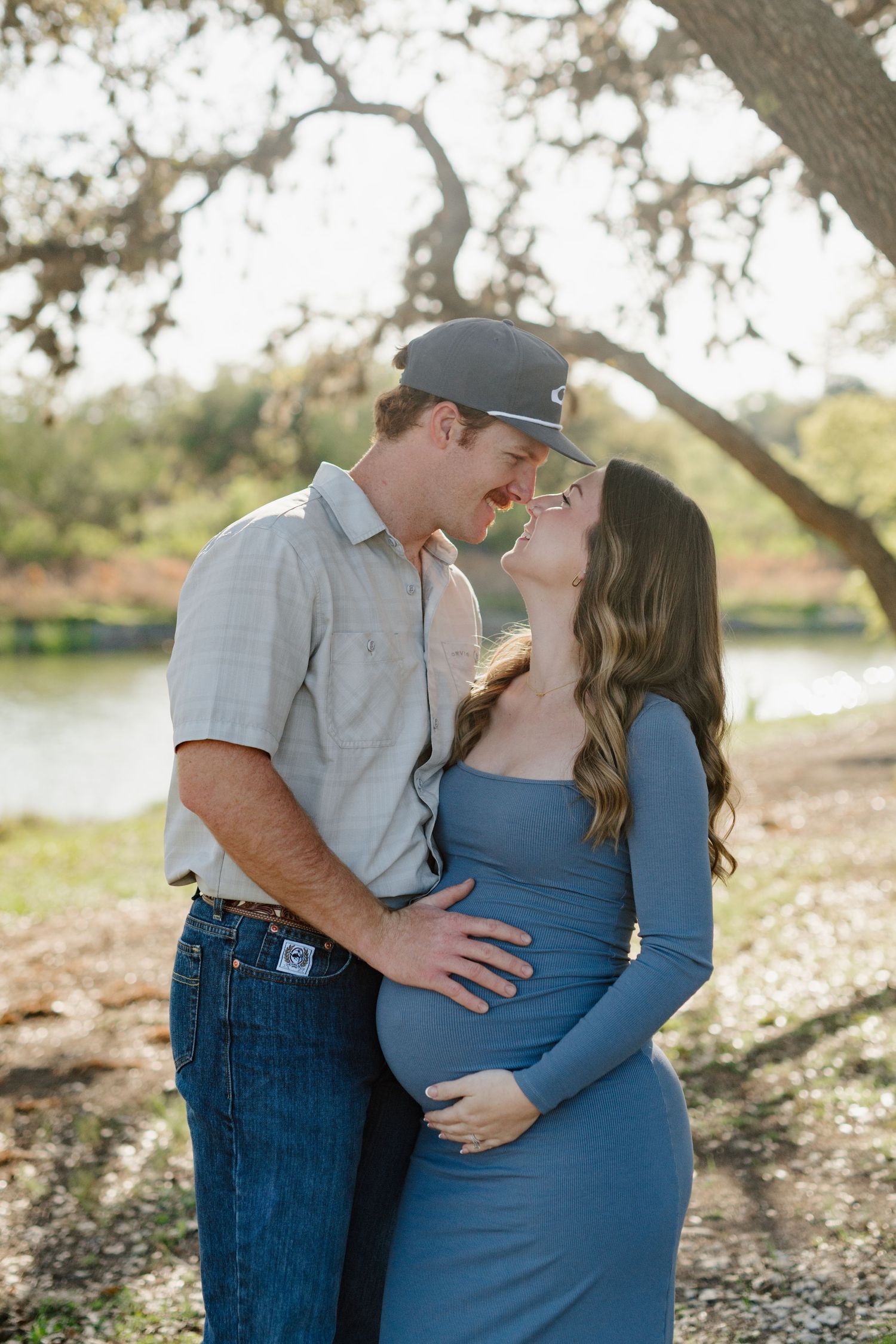 A couple shares a tender moment under a tree by a lake, with her wearing a blue dress while cradling a baby bump.