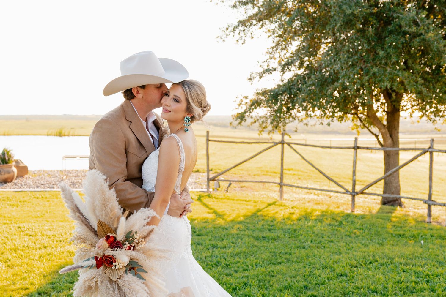 Bride and groom embrace in sunset light on grassy field with fence.