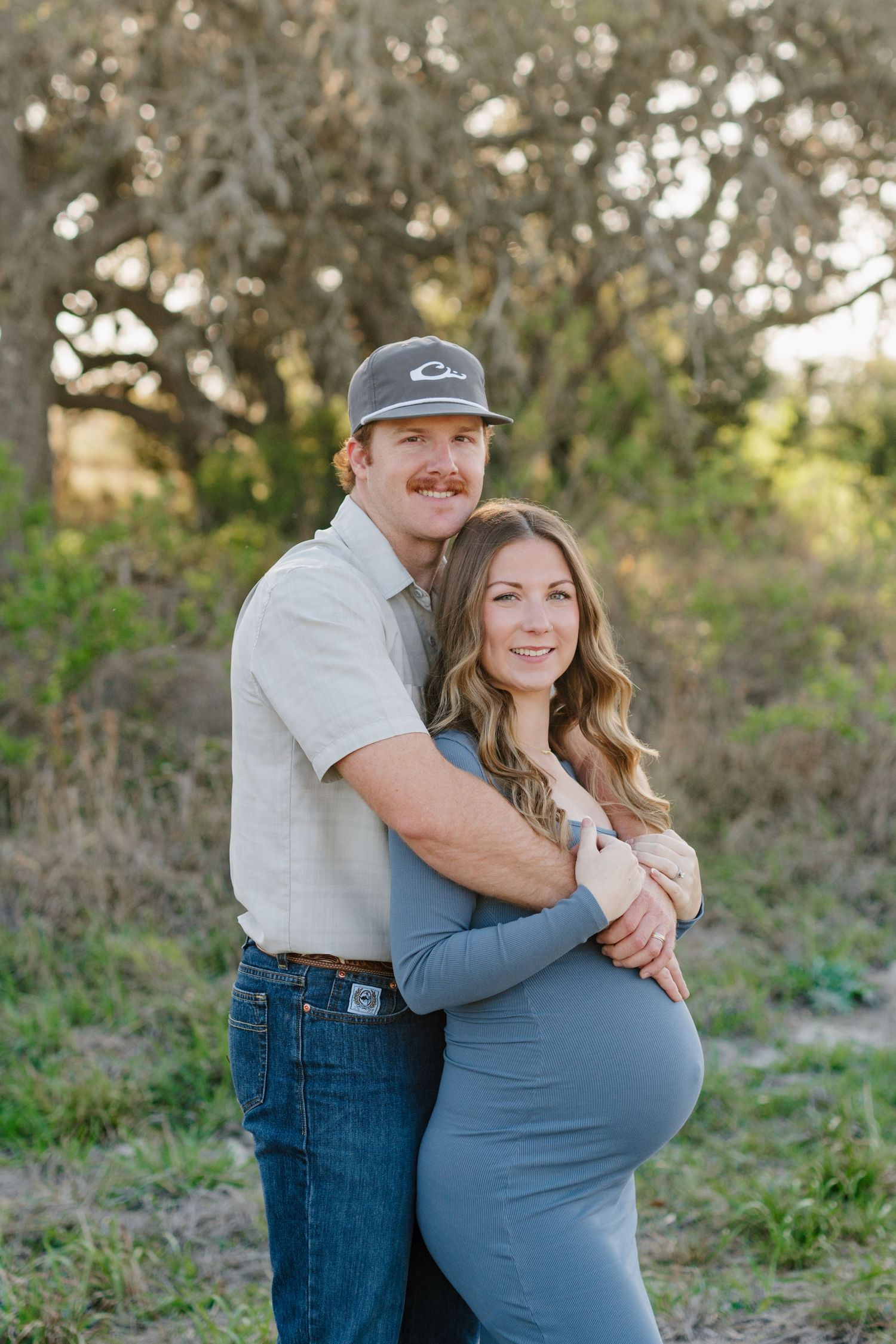 An expectant couple shares a tender embrace outdoors in natural lighting among trees and foliage.