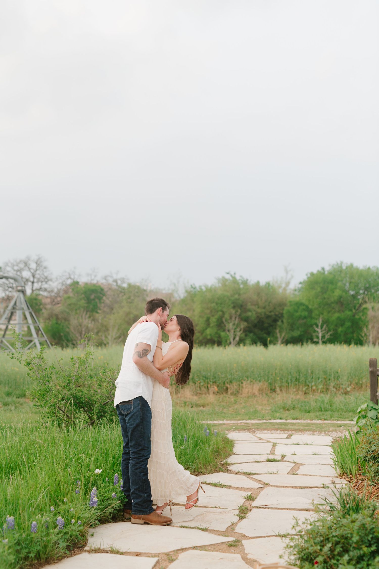 Couple sharing romantic kiss on stone path surrounded by lush greenery and wildflowers.