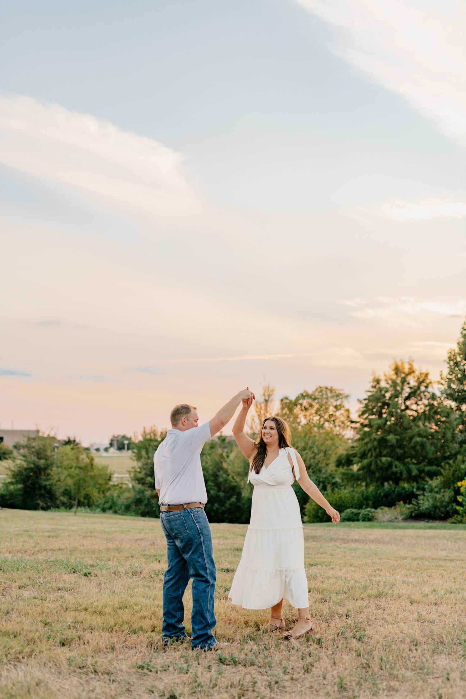 Couple dancing and twirling in a field during golden hour with trees in the background.