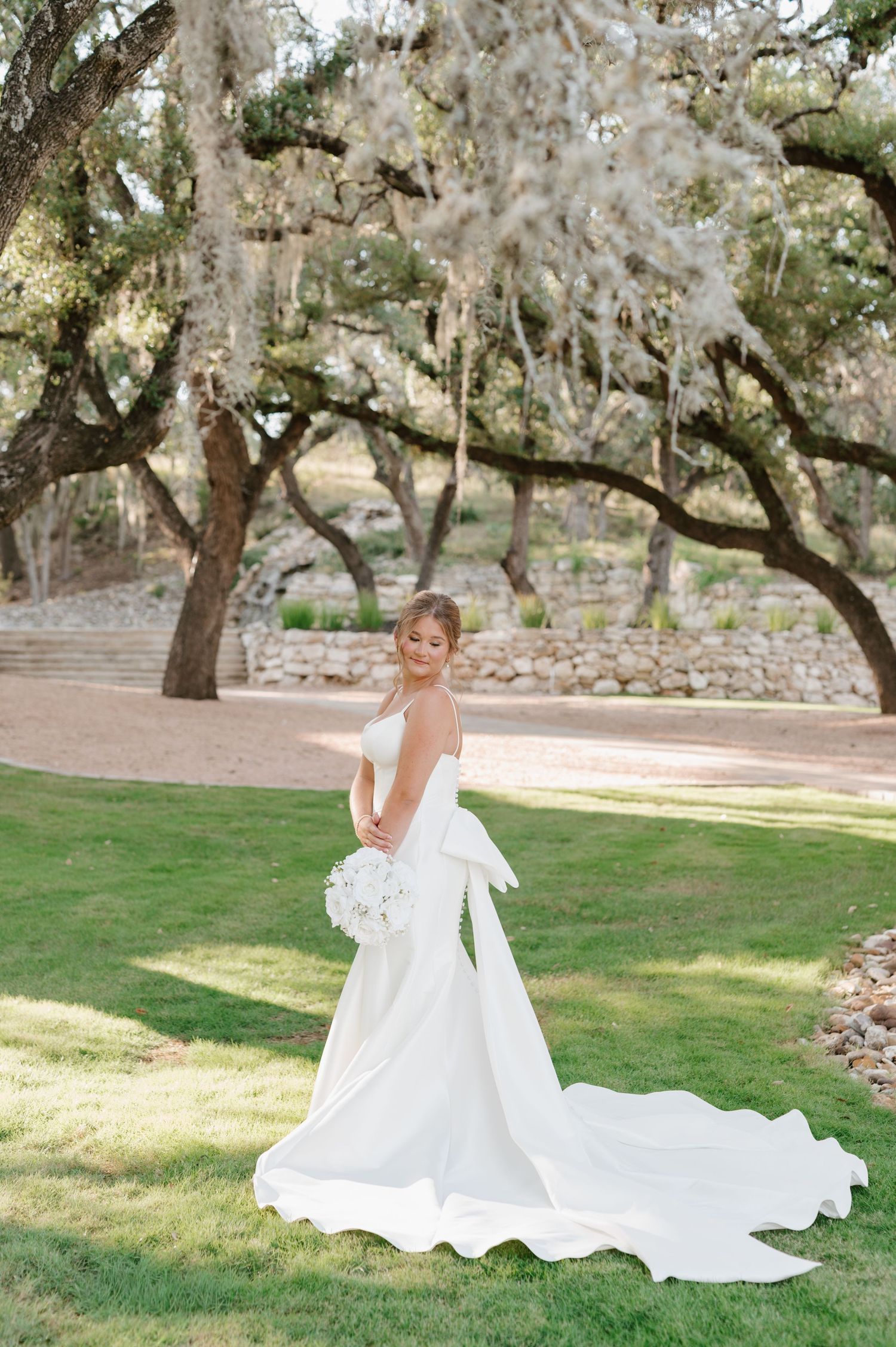 Bride in flowing white gown poses gracefully on grass under Spanish moss-draped trees.
