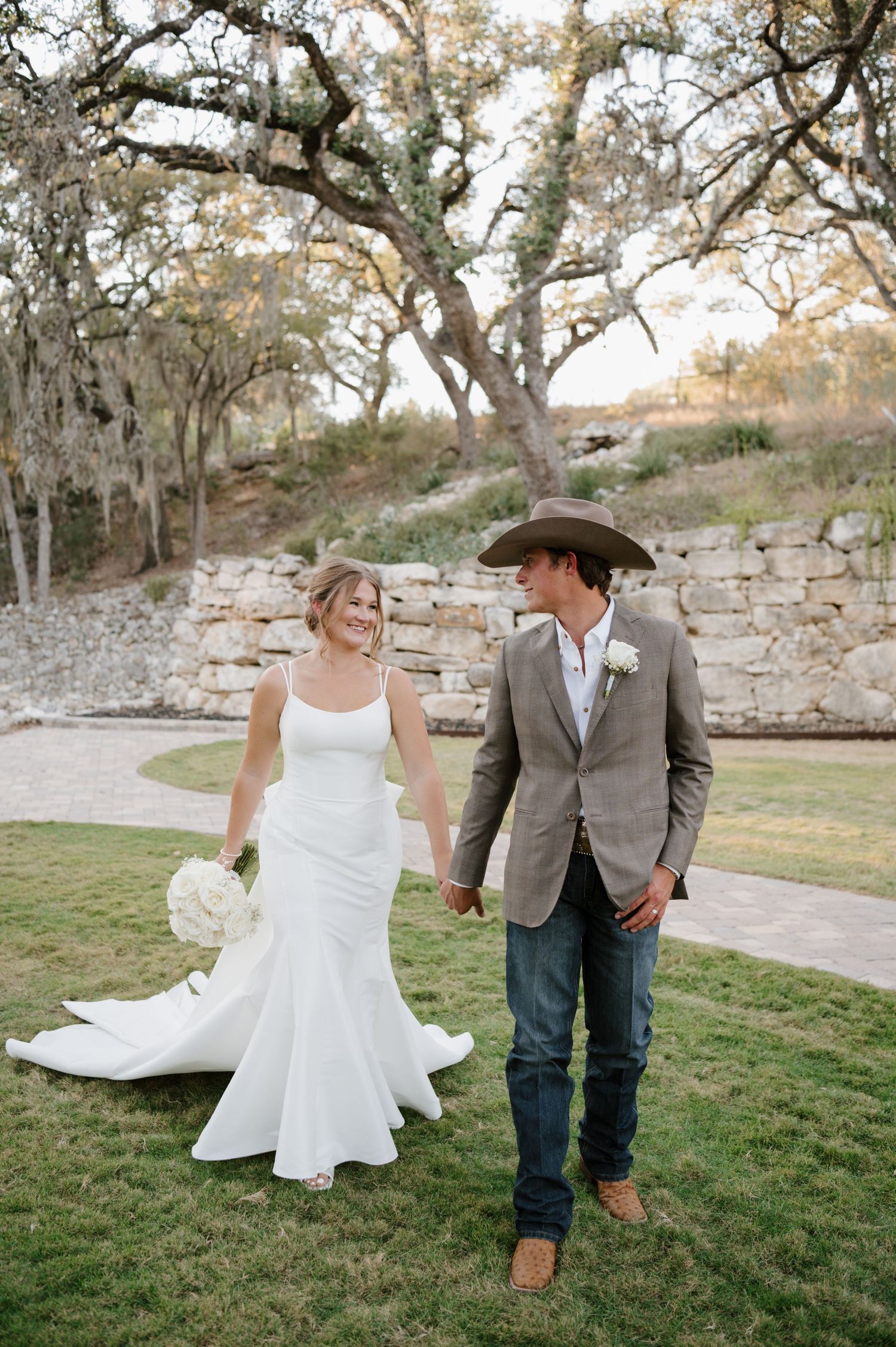 A couple walks hand in hand on their wedding day against a backdrop of oak trees and stone walls.