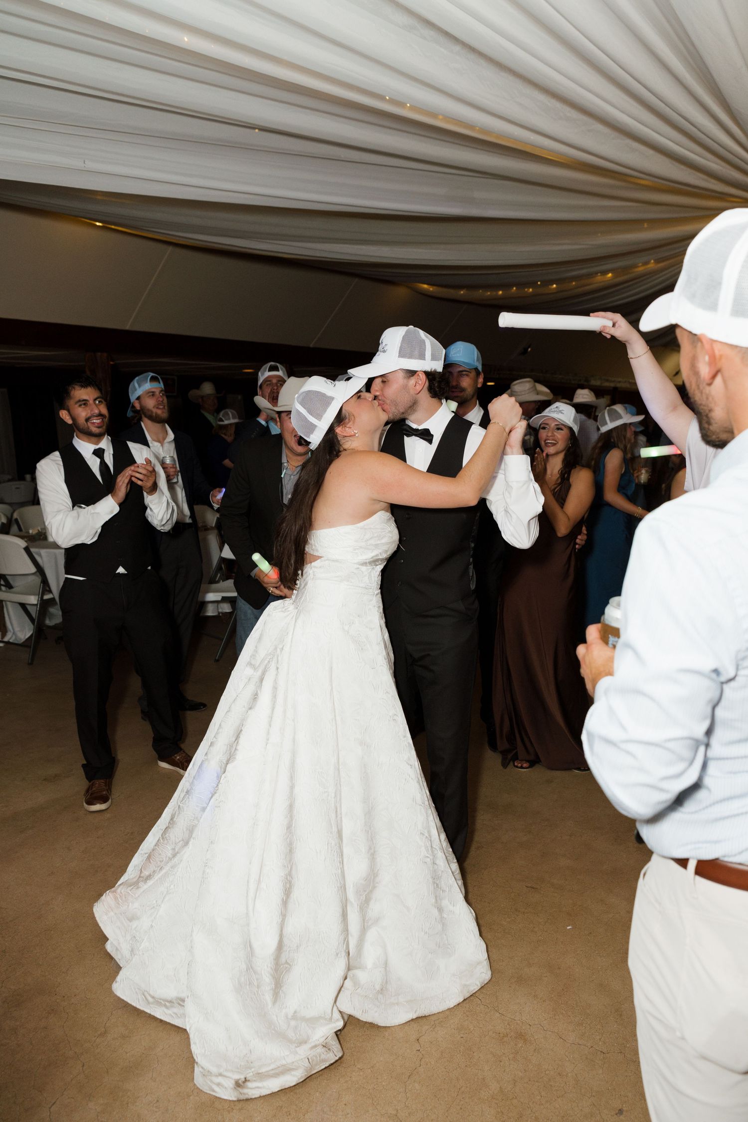 Bride and groom share a kiss on the dance floor while guests wearing white hats celebrate.