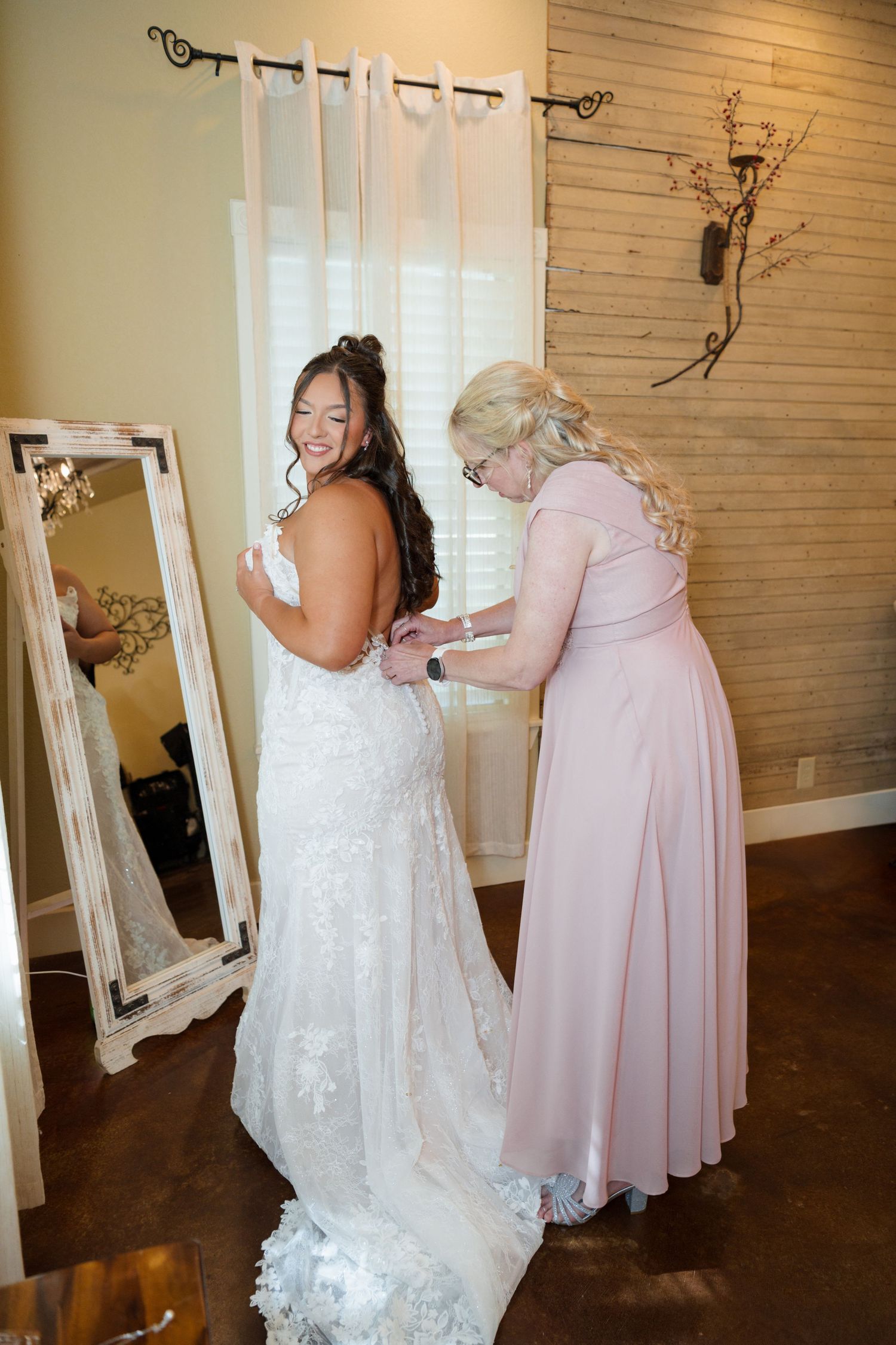 Mother in pink gown buttoning bride's lace wedding dress while bride smiles at mirror.