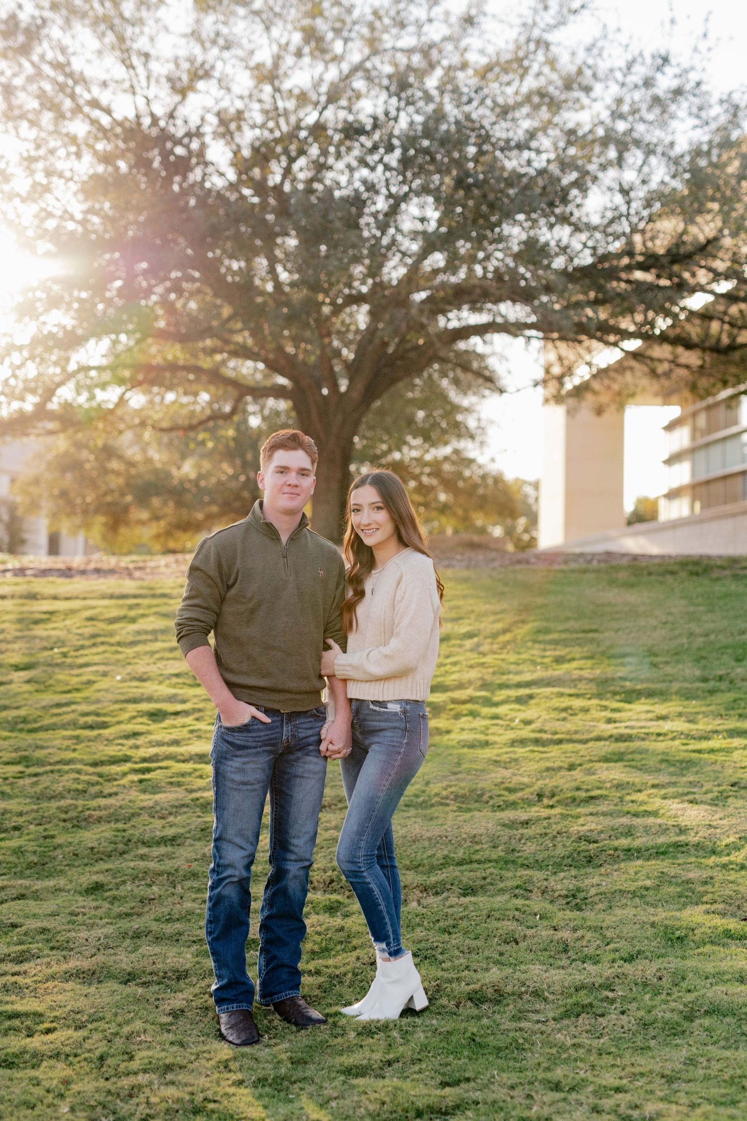 Couple stands together on green grass with a large tree and building in the background.