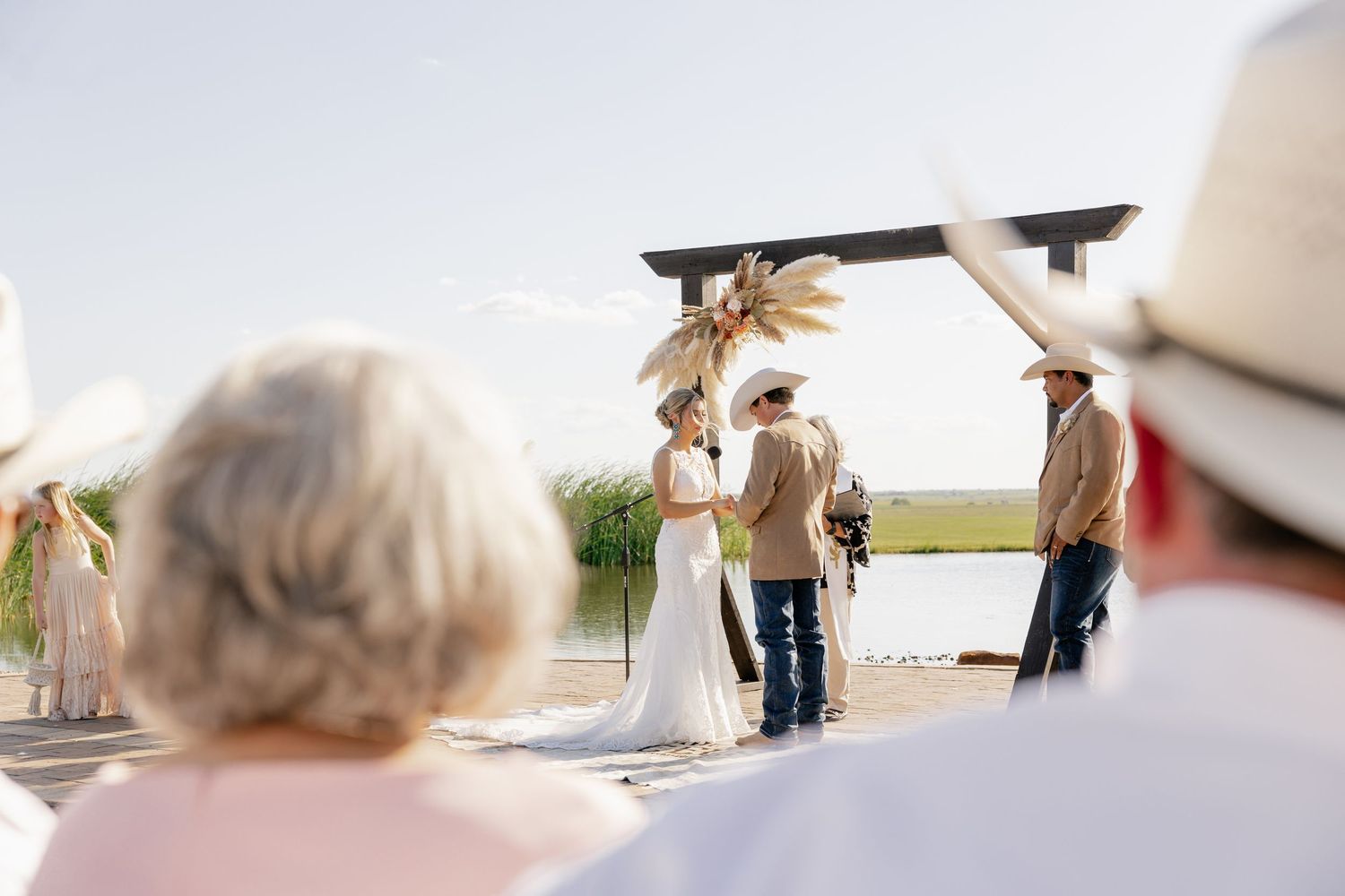 An outdoor wedding ceremony under a wooden arch with guests looking on from white chairs.