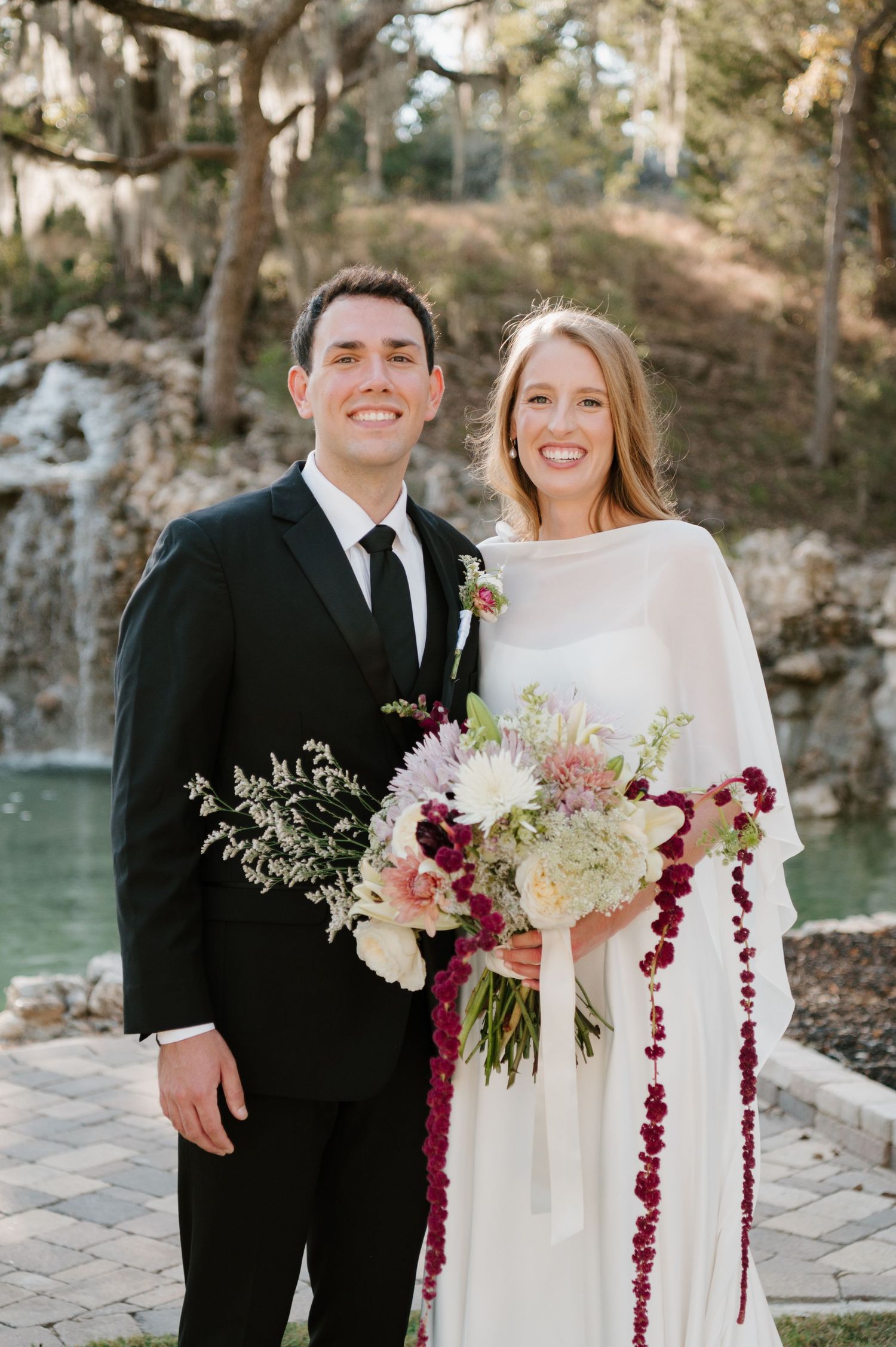 Couple in formal wedding attire poses outdoors with a stunning burgundy and white floral bouquet.