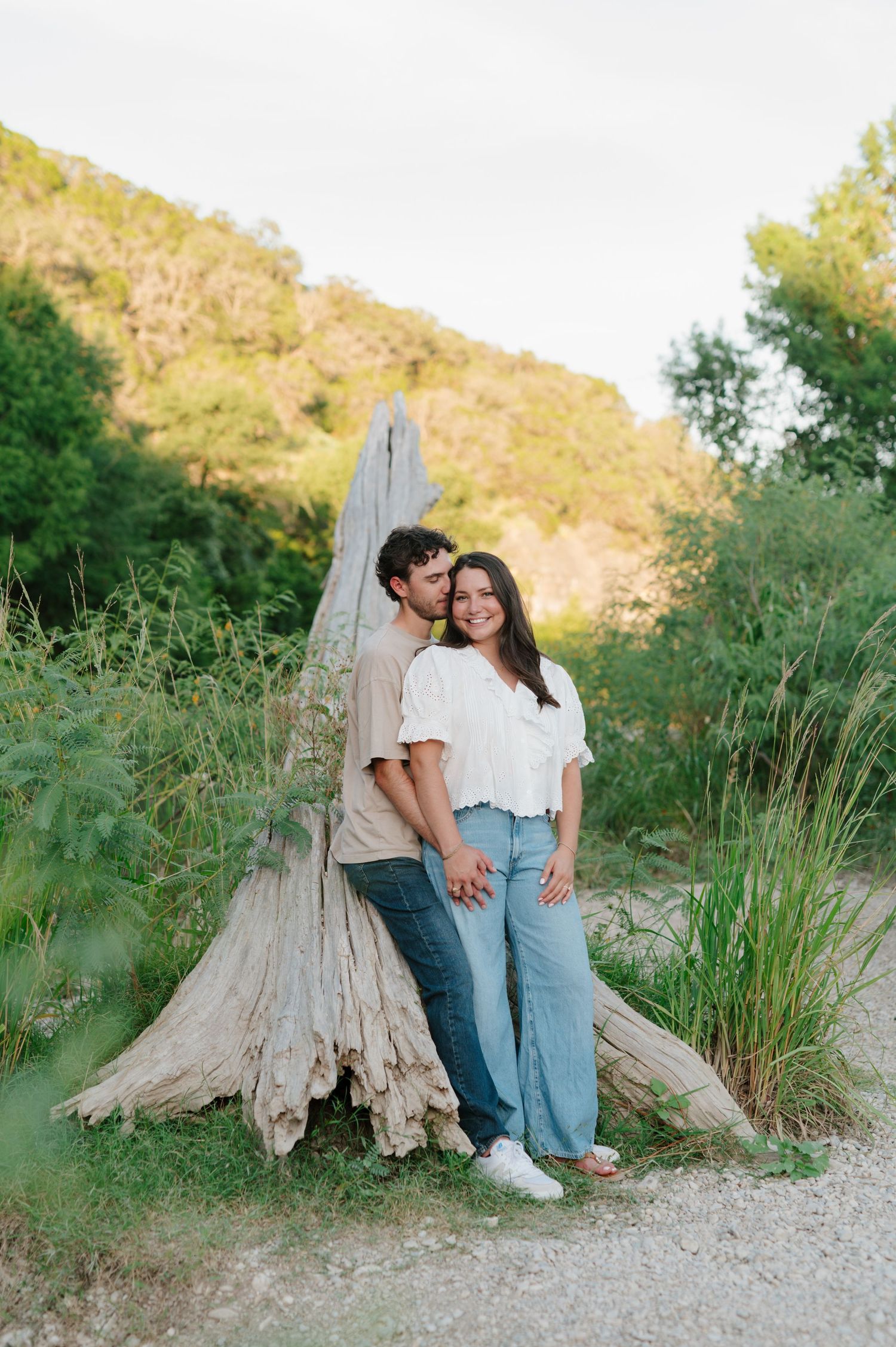 A couple shares an intimate moment near a weathered tree stump in a natural outdoor setting.