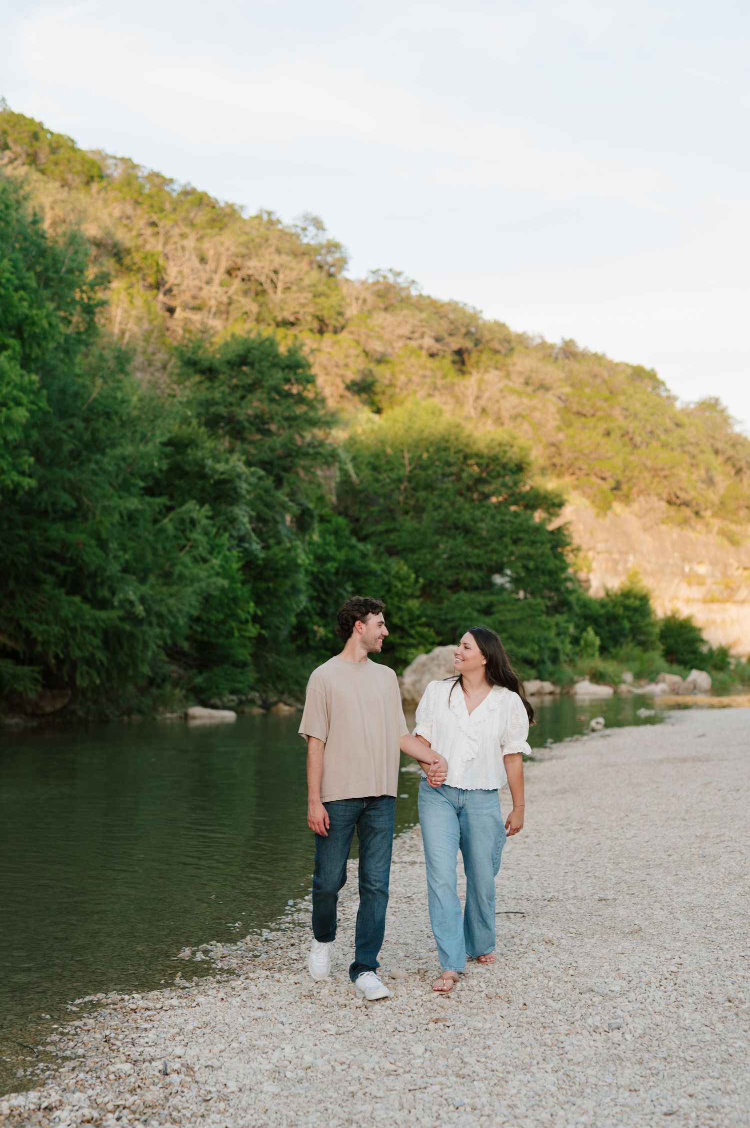 A couple walks together along a scenic riverbank with mountains and trees in the background.