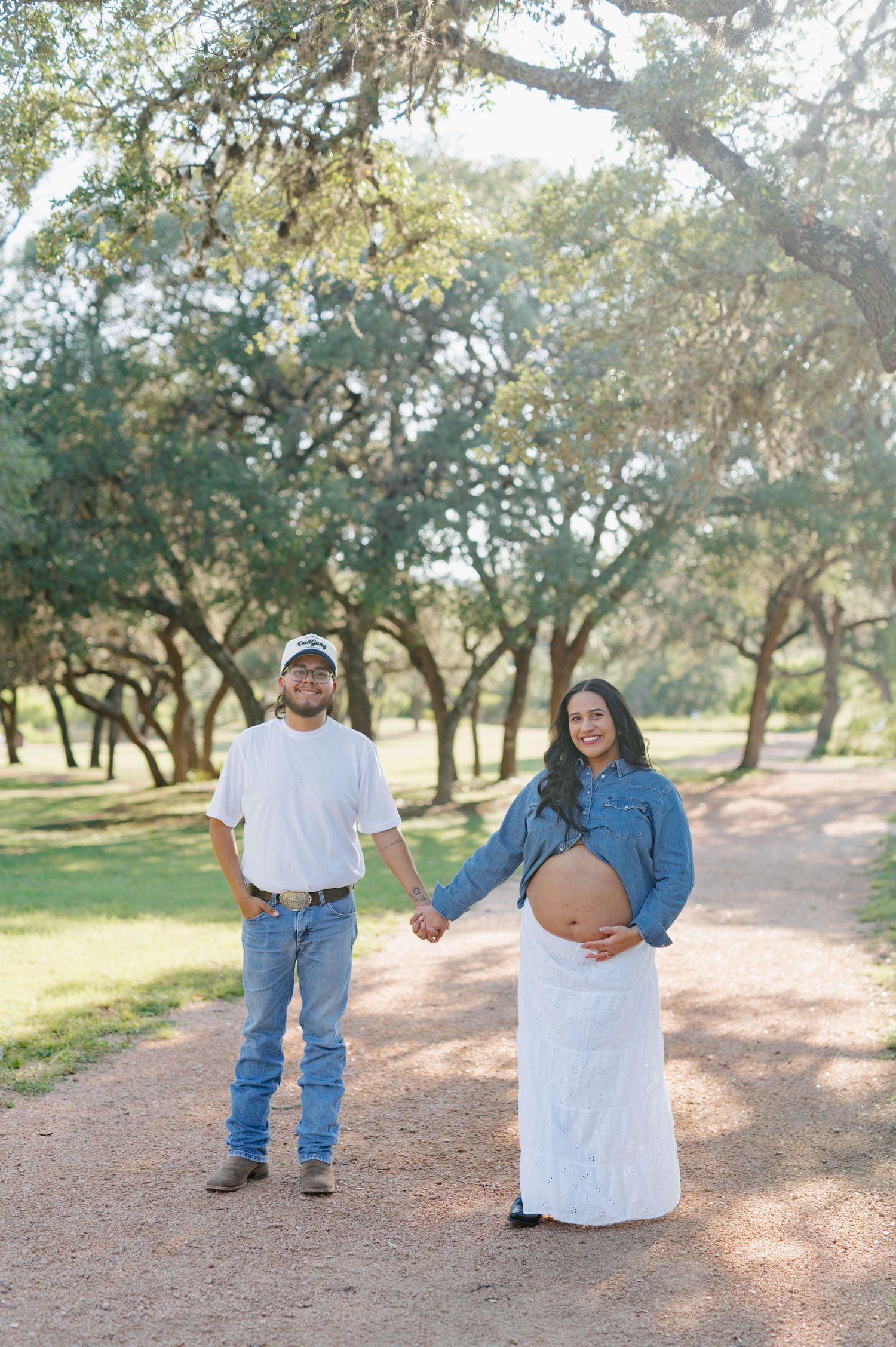 A couple takes romantic maternity photos while walking along a tree-lined dirt path.