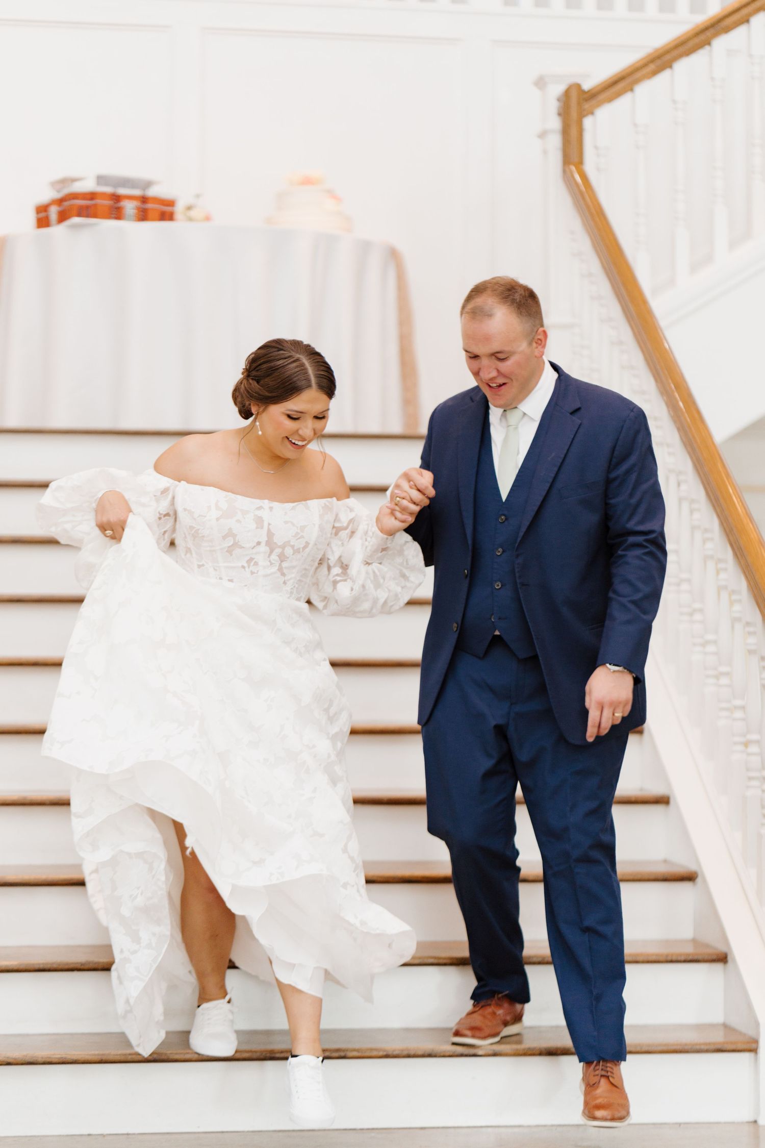 A couple descends white stairs together, with the bride in lace gown and groom in navy suit.