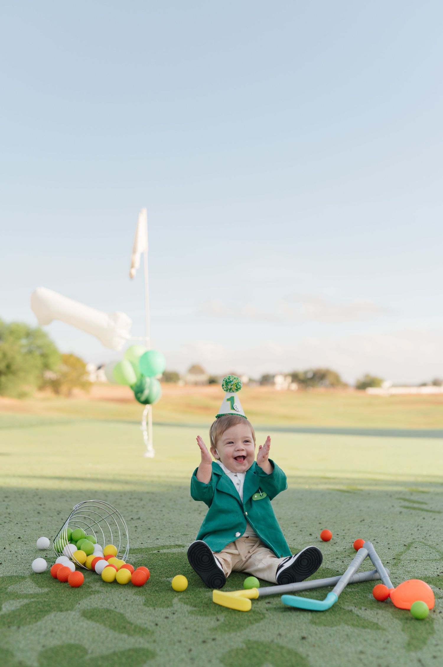Small child in green jacket sits on golf course with colorful golf balls and equipment scattered around.