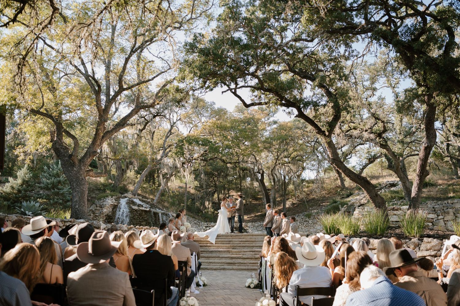 Wedding guests seated on stone steps under oak trees during an outdoor ceremony.