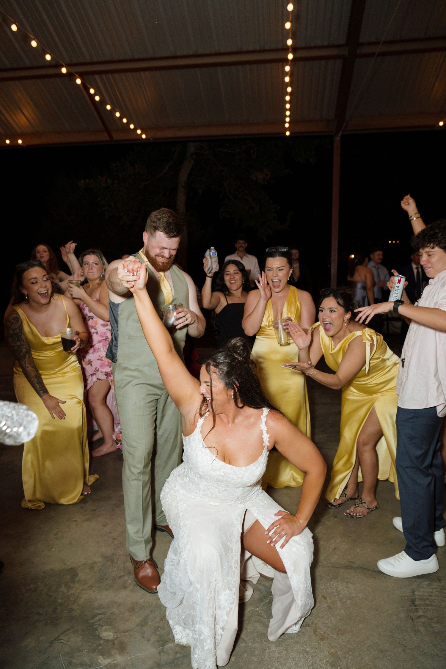 Bride kneels on dance floor removing garter while bridesmaids in yellow dresses cheer at lively outdoor wedding reception.