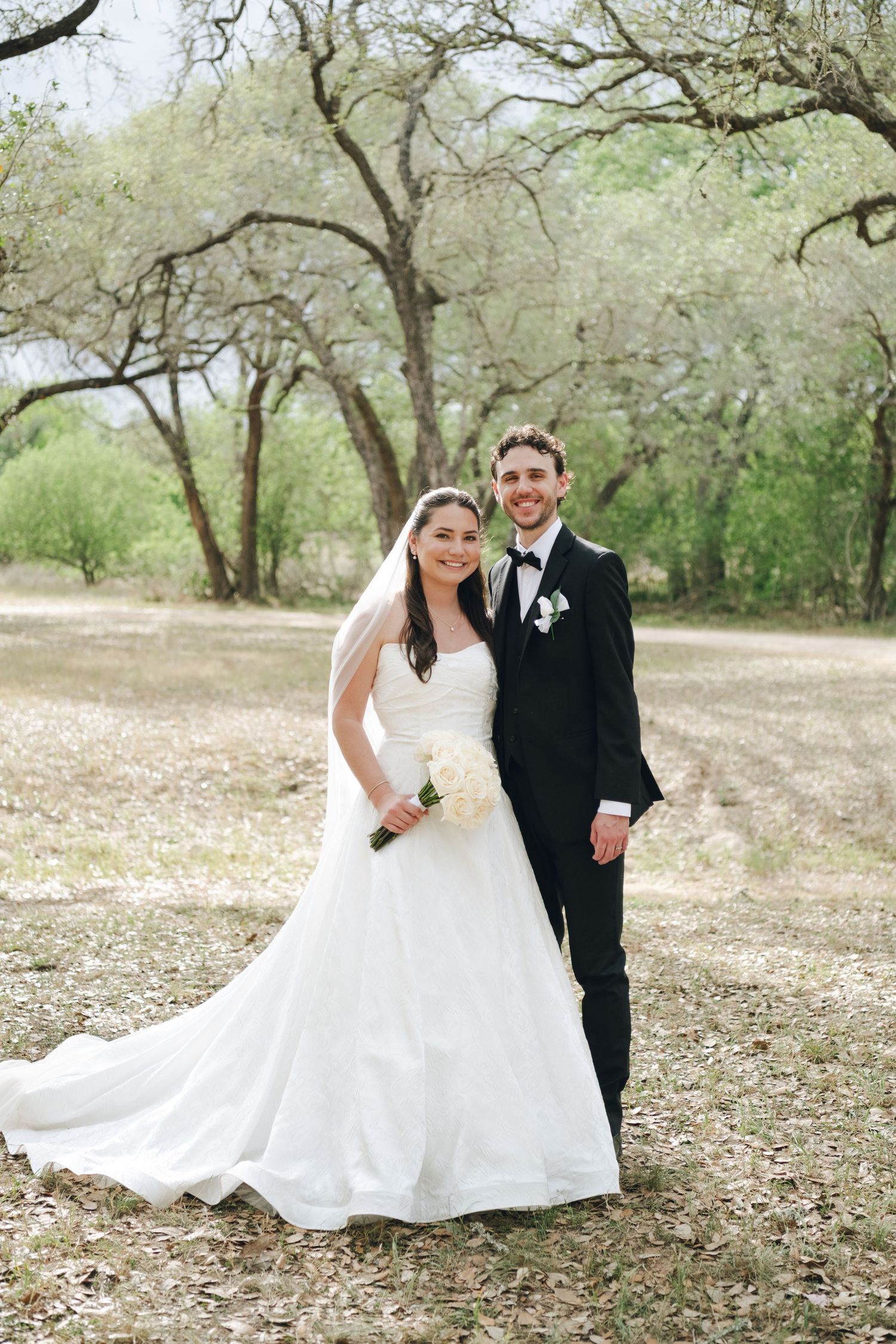 Bride in white gown and groom in black tuxedo pose together outdoors among trees on their wedding day.