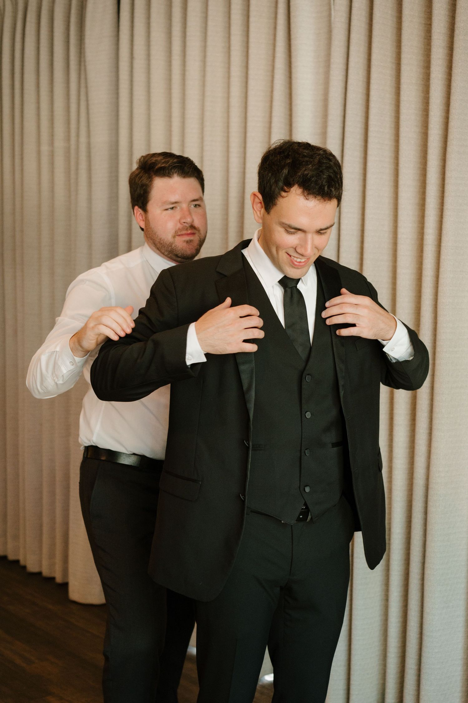 Two men in formal black suits and ties share a lighthearted moment while getting ready for an event.