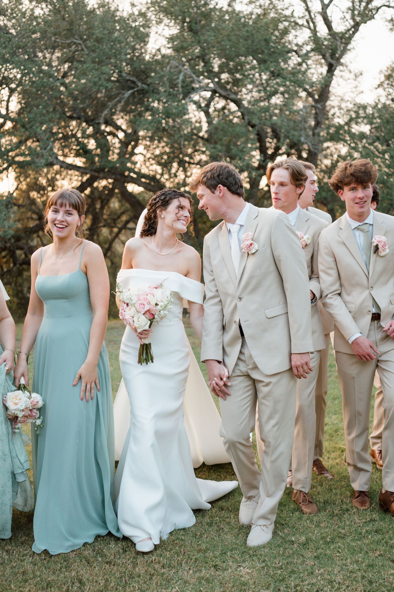 Wedding party in mint green dresses and tan suits poses together on grassy lawn during outdoor celebration.