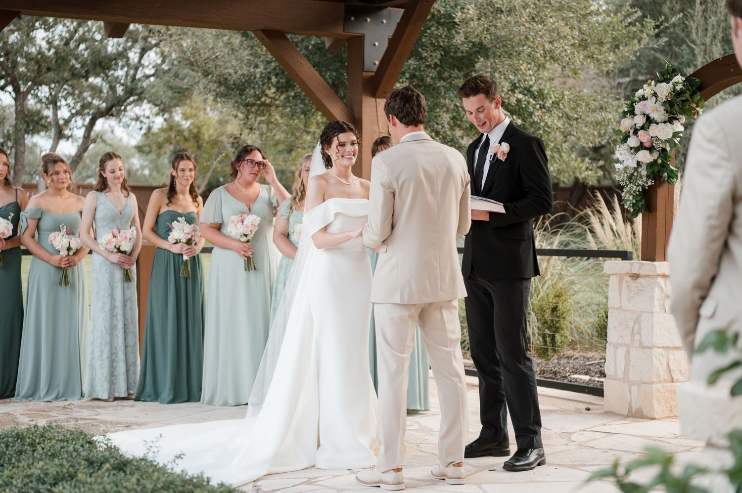 Outdoor wedding ceremony under wooden pergola with bridesmaids in light blue dresses standing to the side.