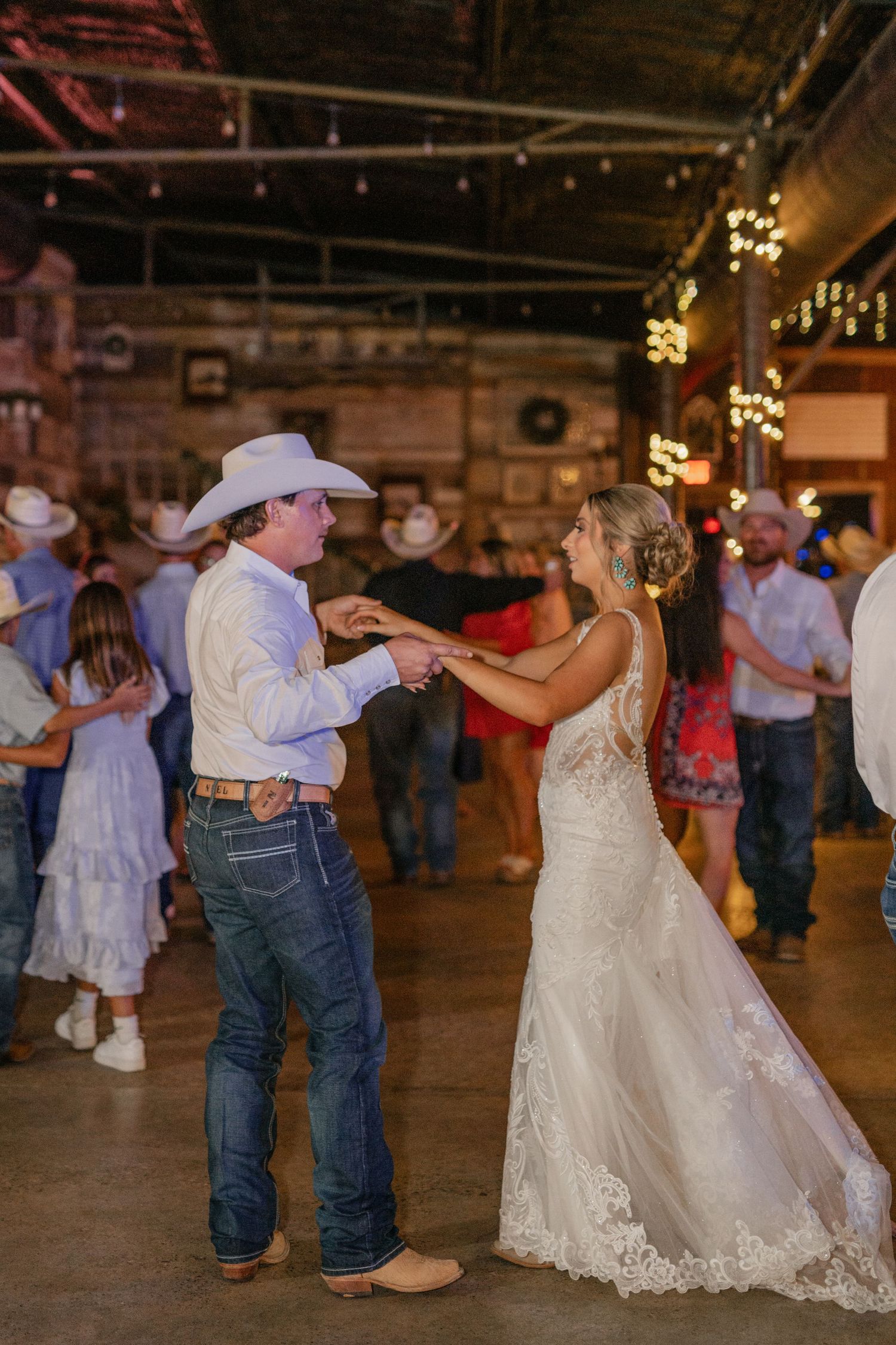 A couple dancing at a rustic barn wedding venue with string lights overhead and guests in the background.