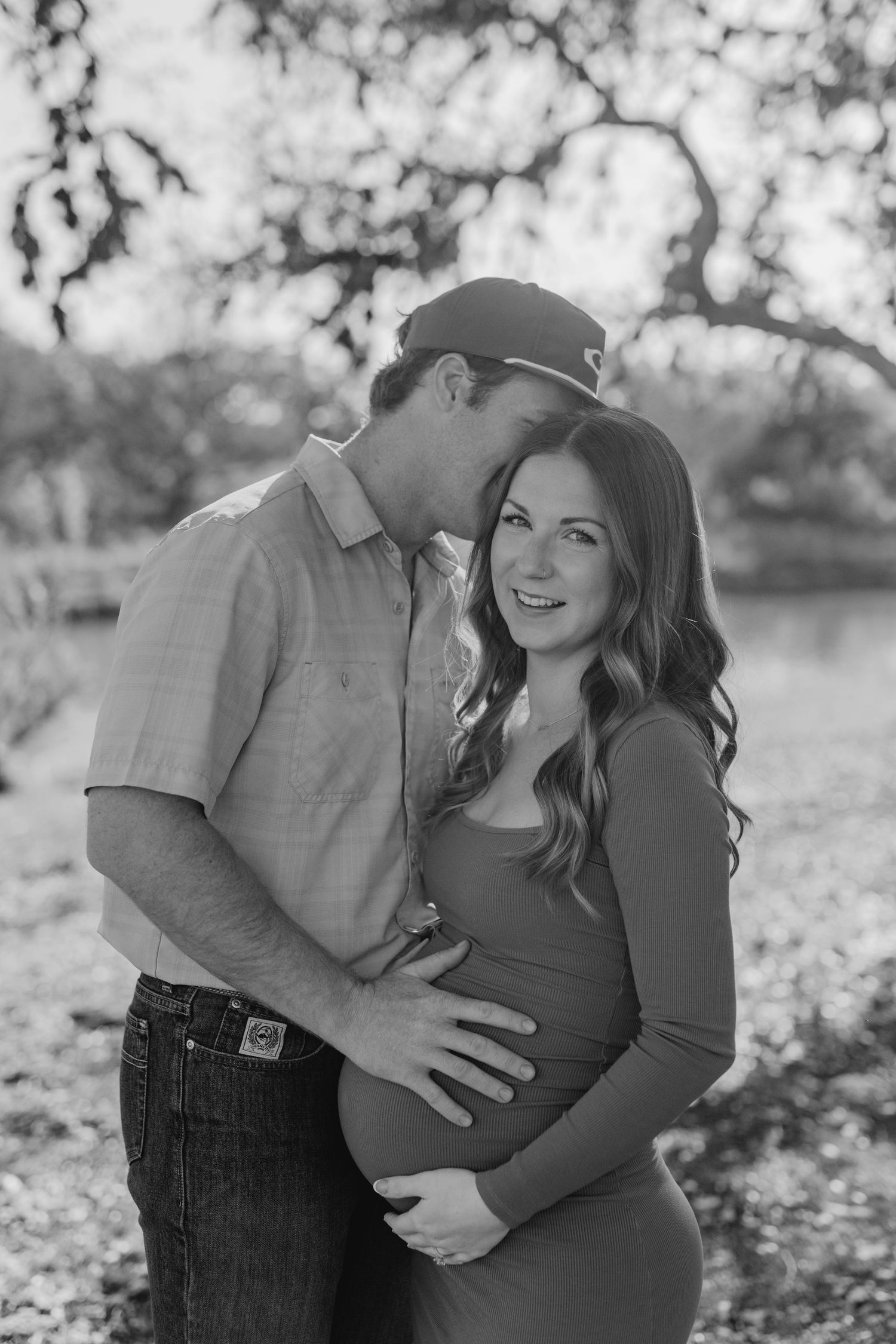 Black and white outdoor maternity photo showing an intimate moment between couple under trees.