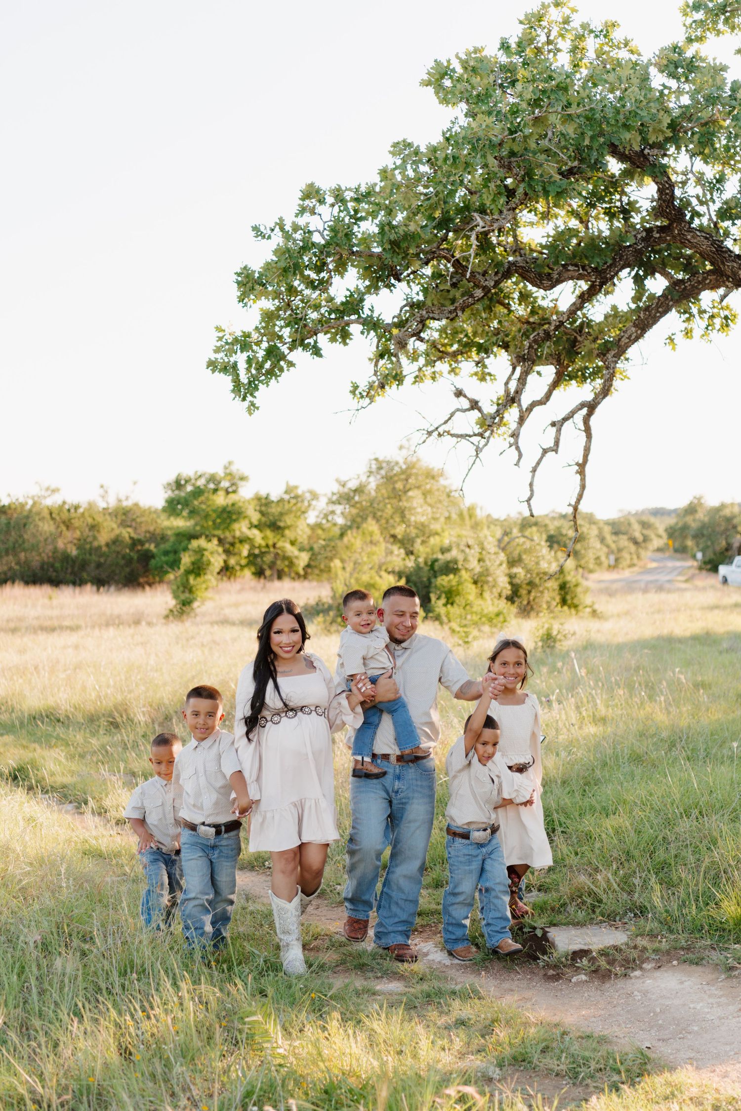 A family poses together on a dirt path beneath a sprawling oak tree in a golden meadow during sunset.