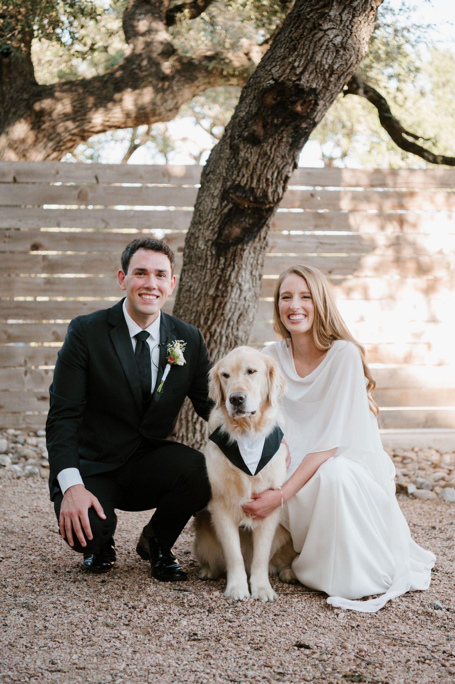 Wedding couple posing outdoors with their golden retriever dog wearing a black bowtie.