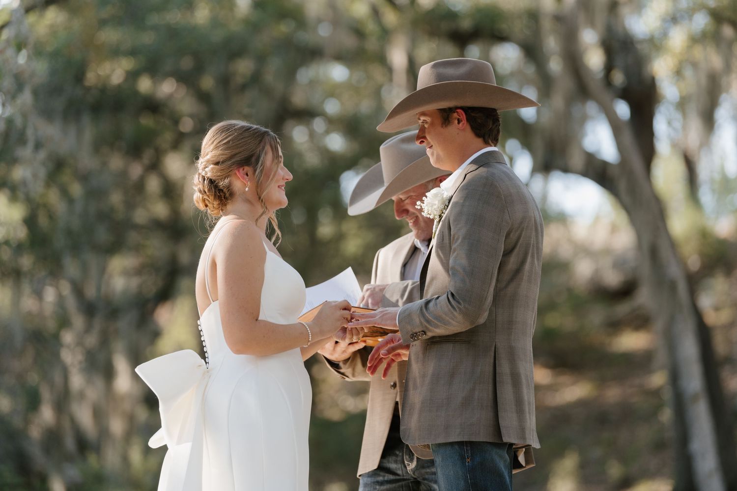 A couple exchanges rings during their outdoor wedding ceremony beneath oak trees.