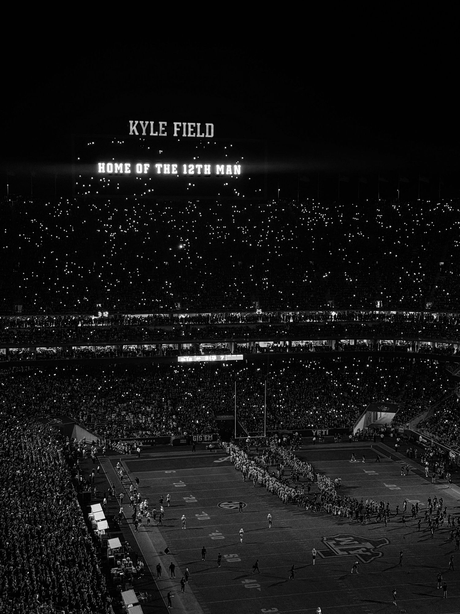 Black and white panoramic view of a crowded football stadium at night with floodlights illuminating the playing field.