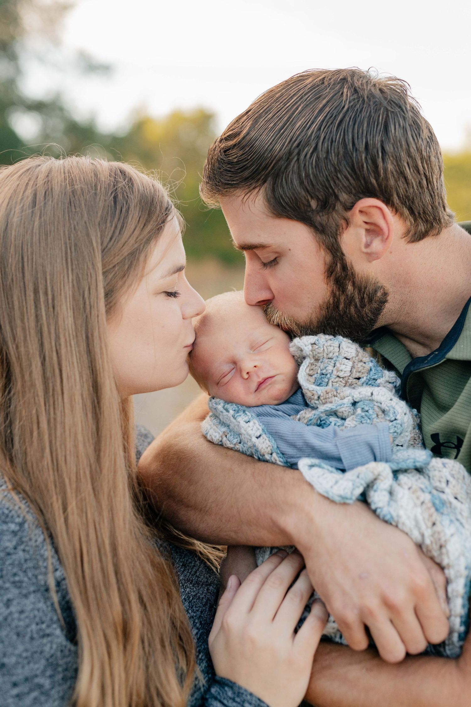 Tender family moment as parents share a loving kiss with their sleeping newborn baby in blue outfit.
