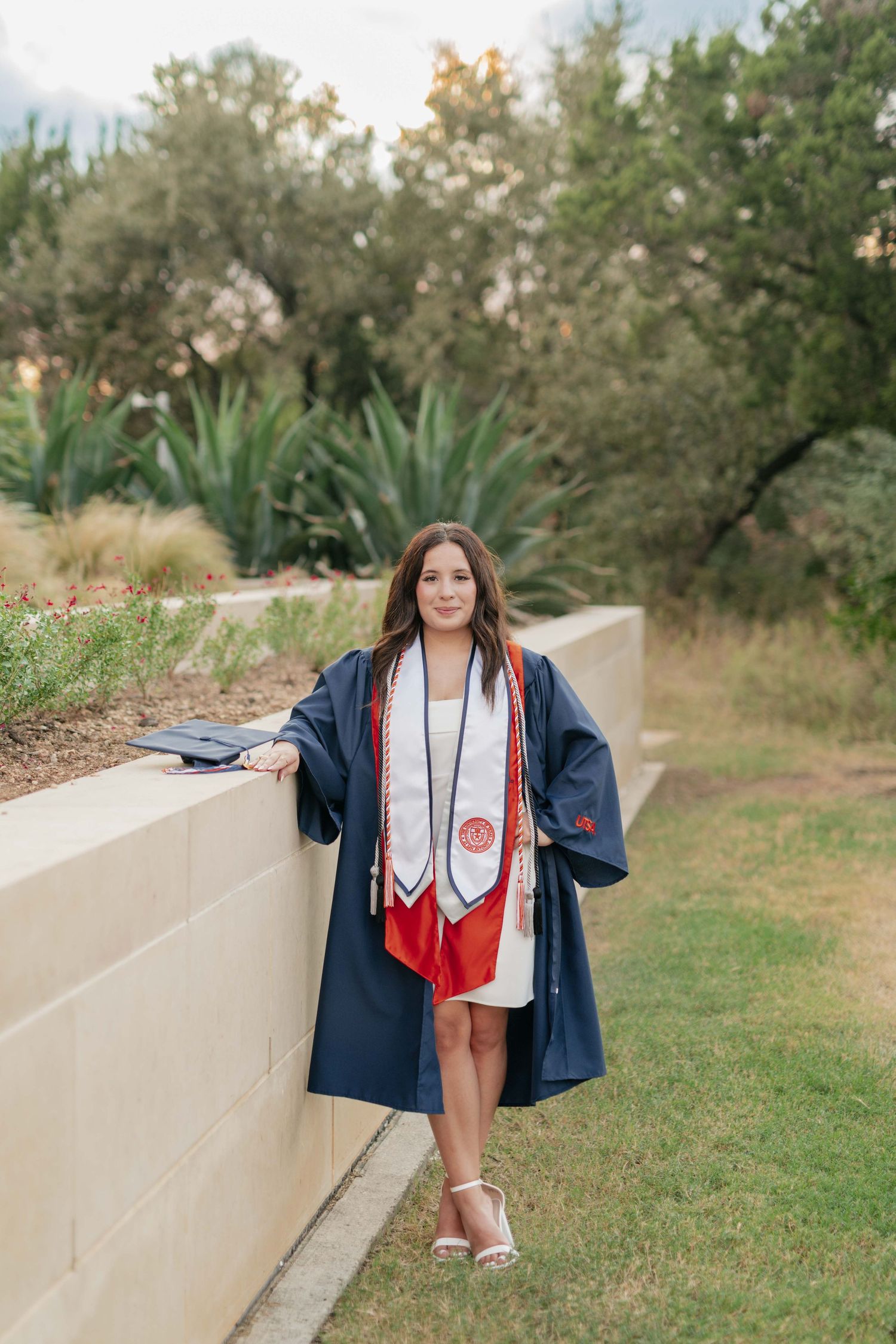 Graduate in navy cap and gown with red stole poses by landscaped wall outdoors.