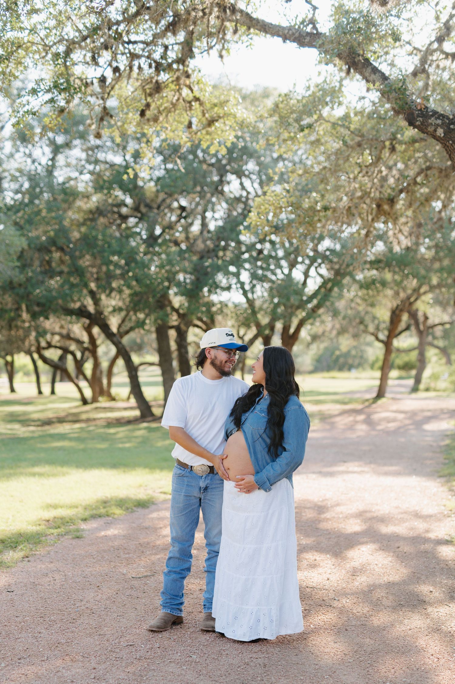 A romantic couple poses together in a sun-dappled grove of oak trees on a dirt path.
