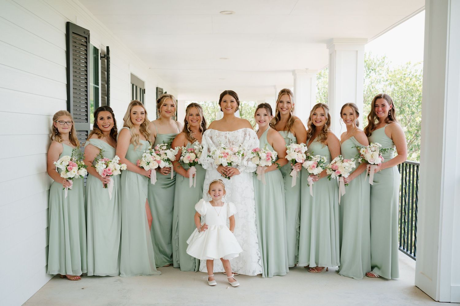 Wedding party wearing sage green dresses standing in a bright white venue.