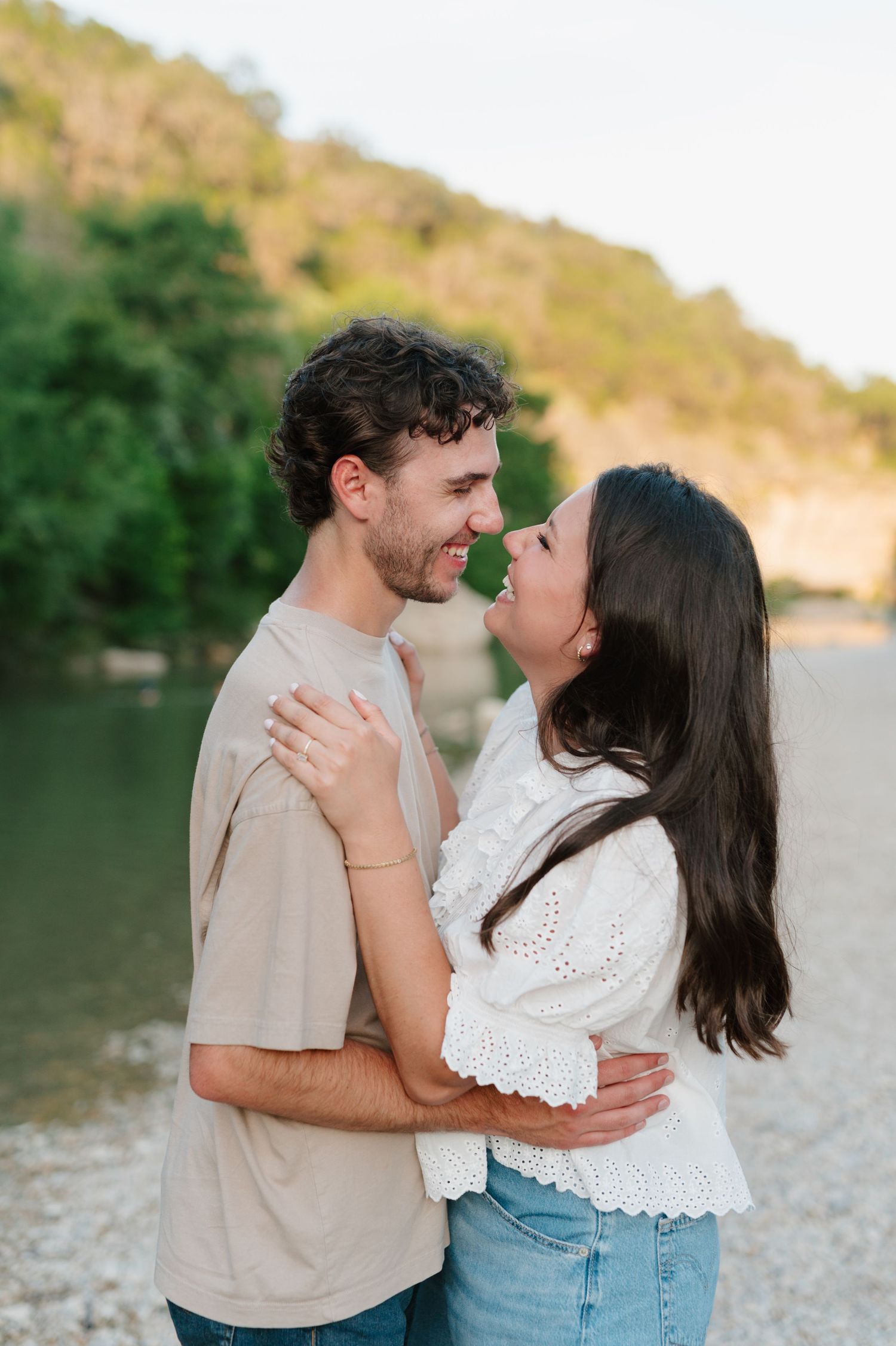 Intimate moments captured between a couple in casual attire by a riverside with natural scenery.