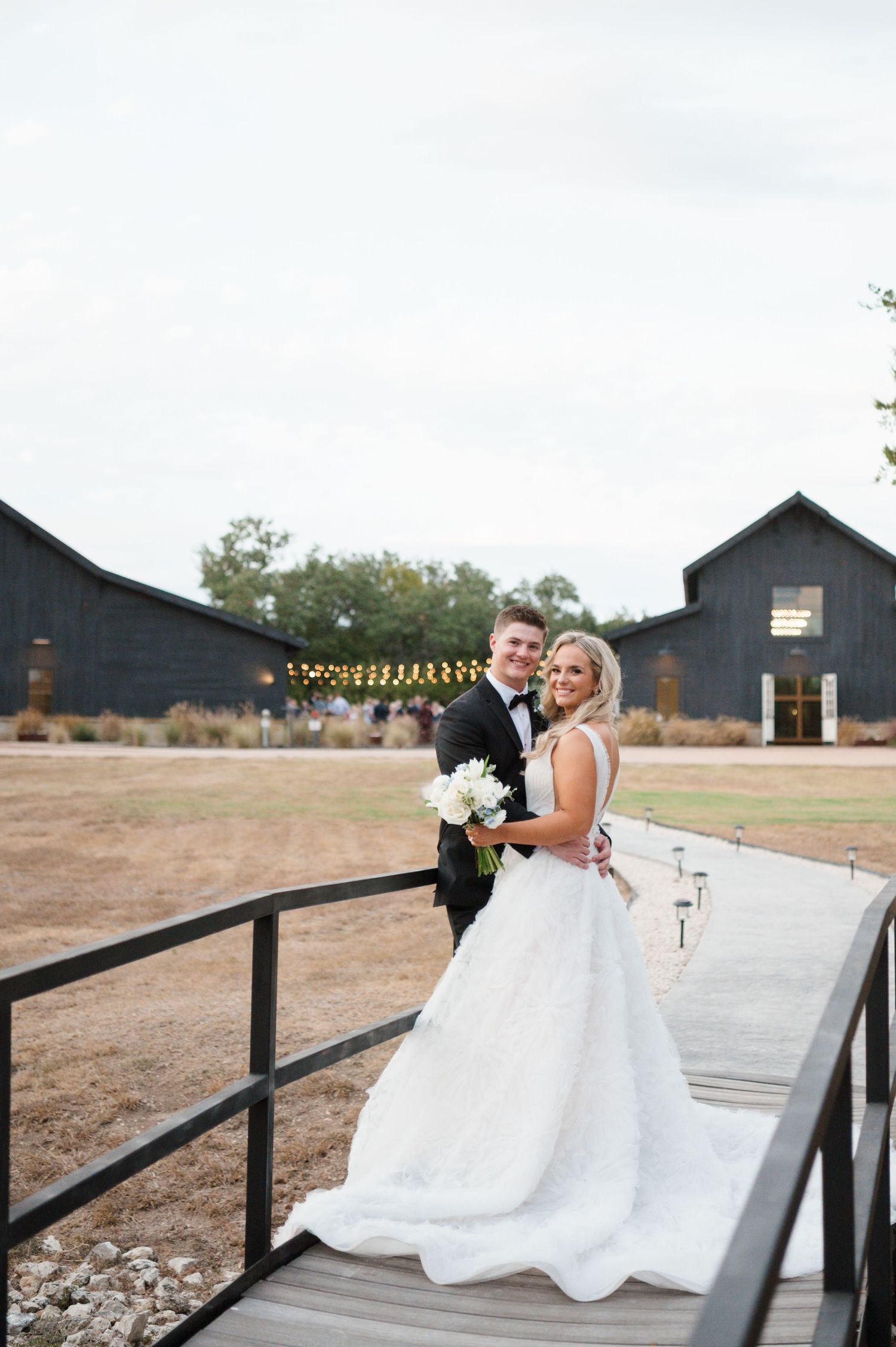 A bride and groom pose together on a wooden bridge in a rustic outdoor setting with dark buildings in the background.