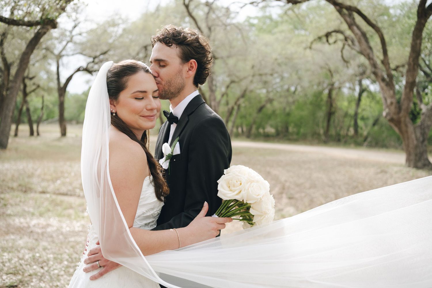 Groom tenderly kisses bride on the cheek as she holds a white rose bouquet outdoors.