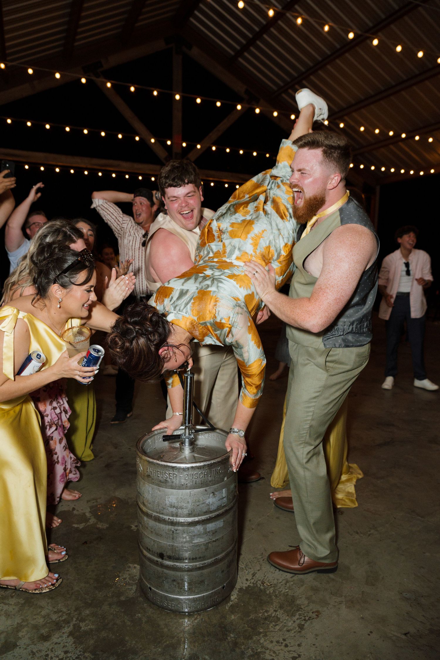 A lively wedding reception scene with guests dancing as a man lifts a woman onto a barrel indoors.