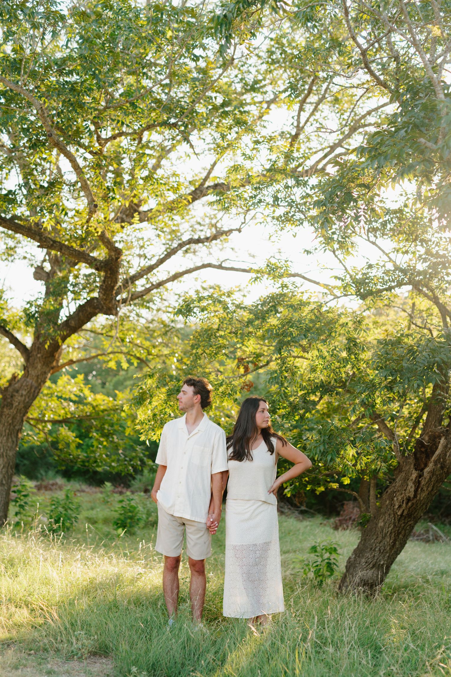 Couple in white clothing shares tender moments under trees with golden sunlight filtering through.