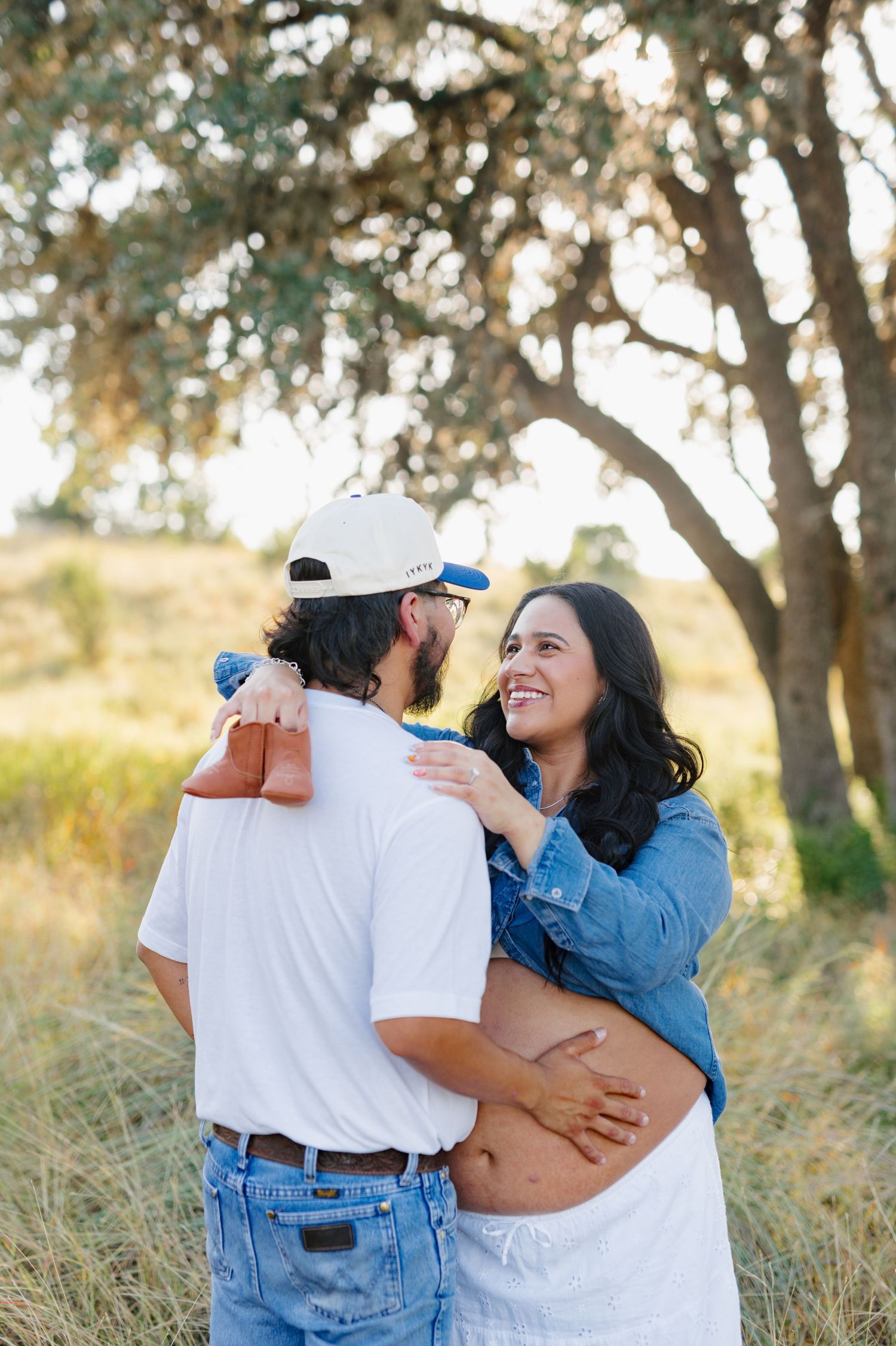 An expecting couple shares intimate embrace in a sunny outdoor setting with trees.